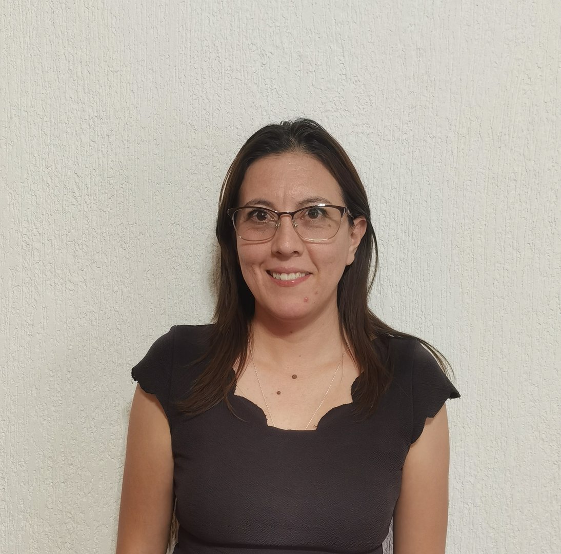 A woman with glasses and long hair wearing a dark top, smiling in front of a plain white textured wall.