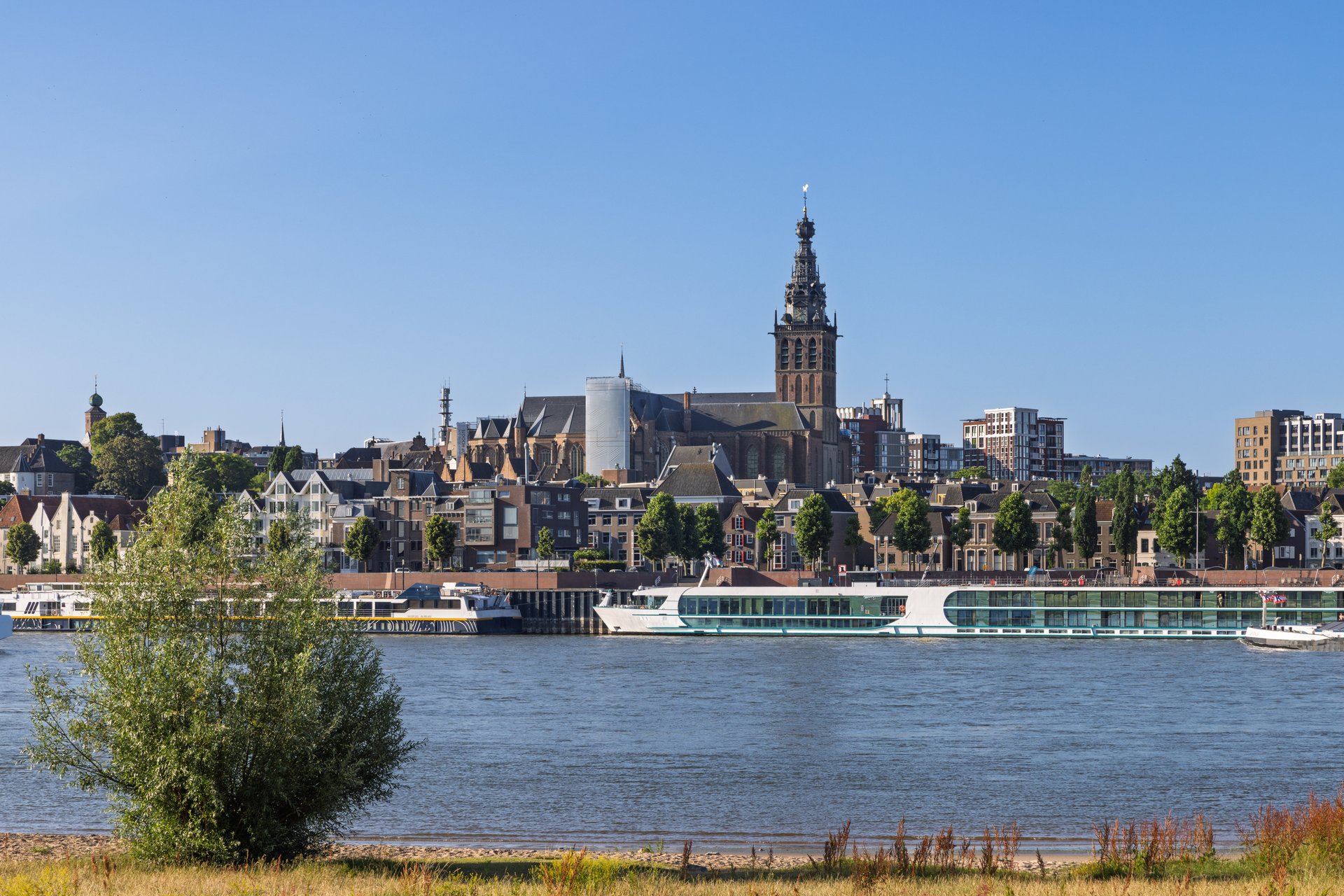 The magnificent Stevenskerk church spire dominates the historic skyline of Nijmegen, Netherlands, as river cruise ships dock along the Waal, all bathed in clear summer sunlight. Nijmegen, Netherlands, 20 May 2025.