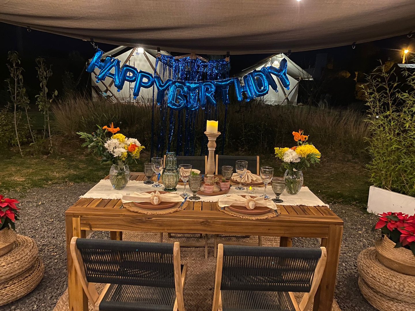 Outdoor birthday dinner setup with wooden table, flowers, plates, candles, and a blue Happy Birthday banner in the background.