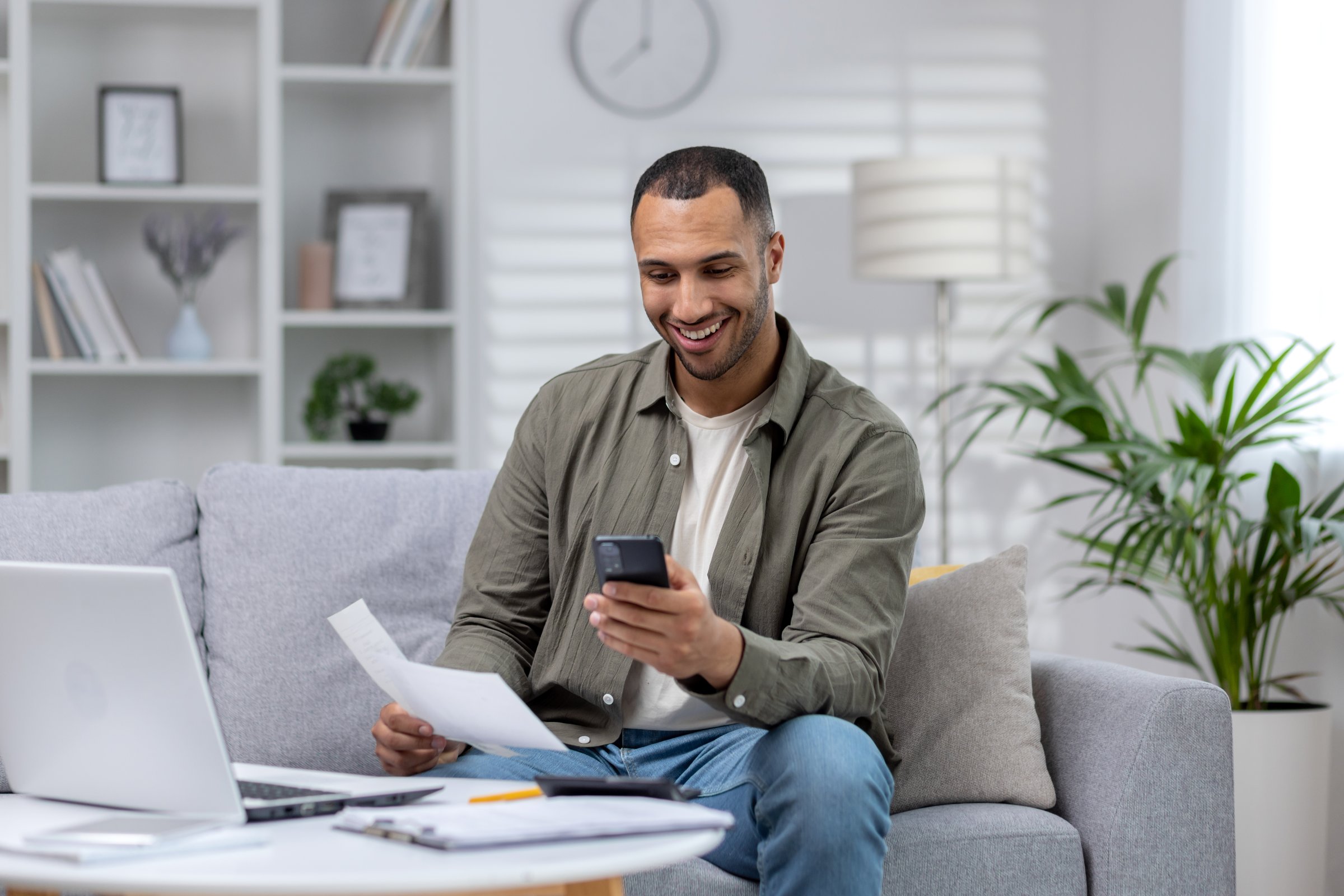 Young smiling African-American man sitting on sofa at home, holding phone and documents, working on laptop.