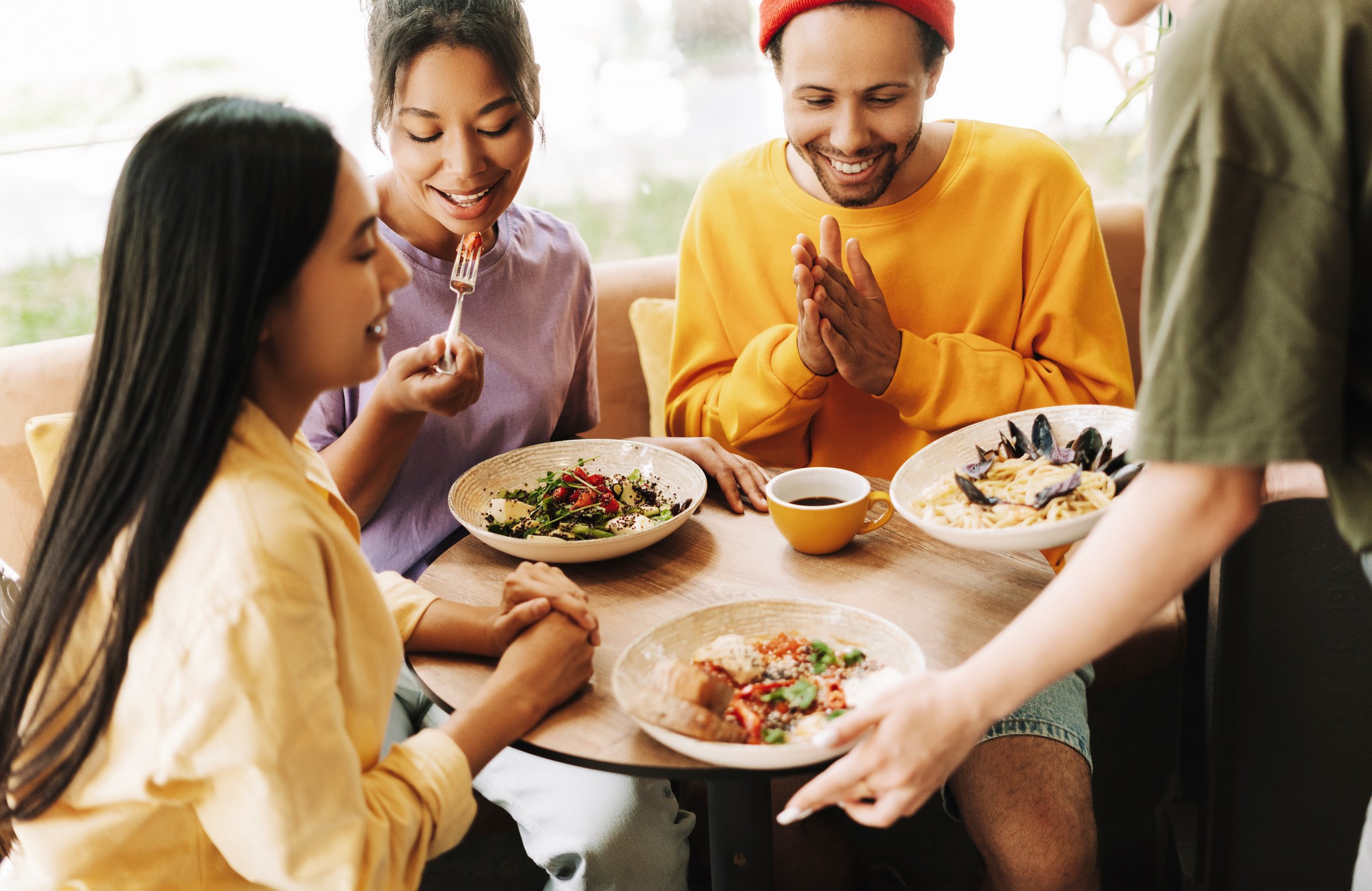 Customers enjoying a meal in a modern cafe, chatting happily. A diverse group of friends sharing salads and pasta, with natural light streaming in