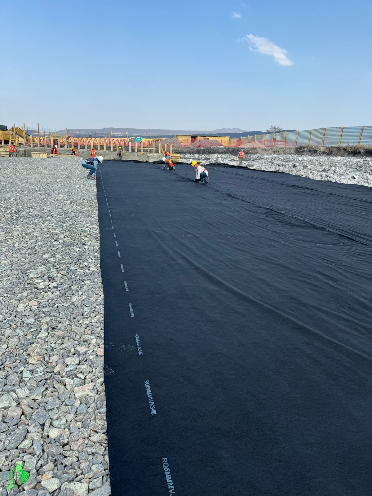Workers installing a large black geotextile sheet over a rocky surface at a construction site under a clear sky.