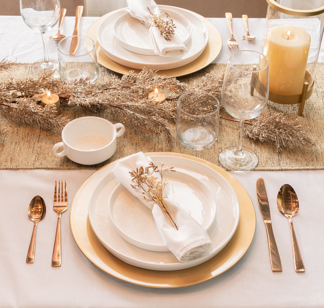 Elegant and luxurious table setting for a festive Christmas or holiday dinner. The setup uses a white tablecloth, gold charger plates, white dishes, sparkling gold tinsel, and rose gold flatware, illuminated by warm candlelight. This high-angle view captures the upscale, celebratory mood of the dining experience.