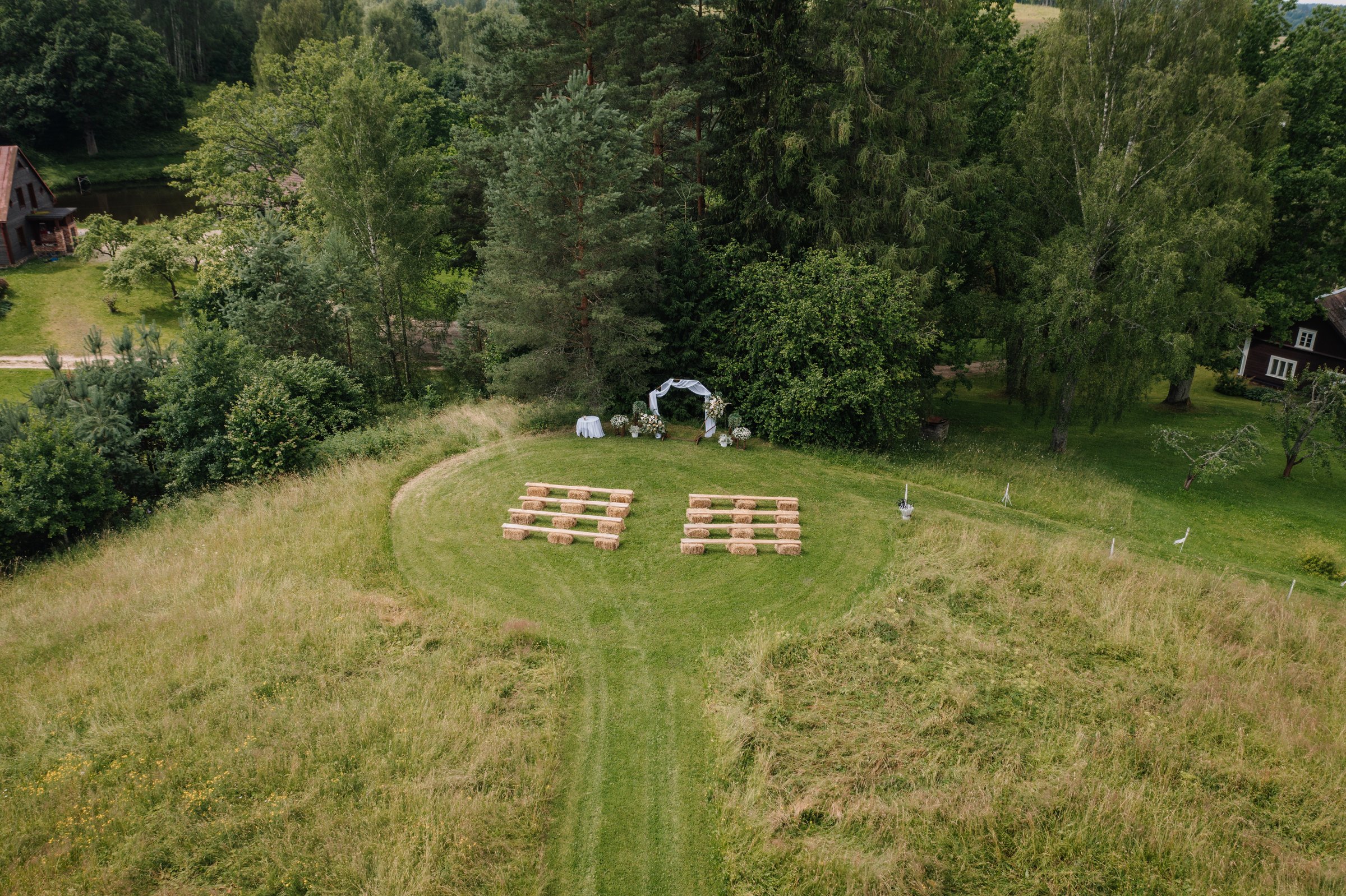 Aerial view of an outdoor countryside wedding ceremony setup with hay bale seating and floral arch, surrounded by trees and rustic buildings.