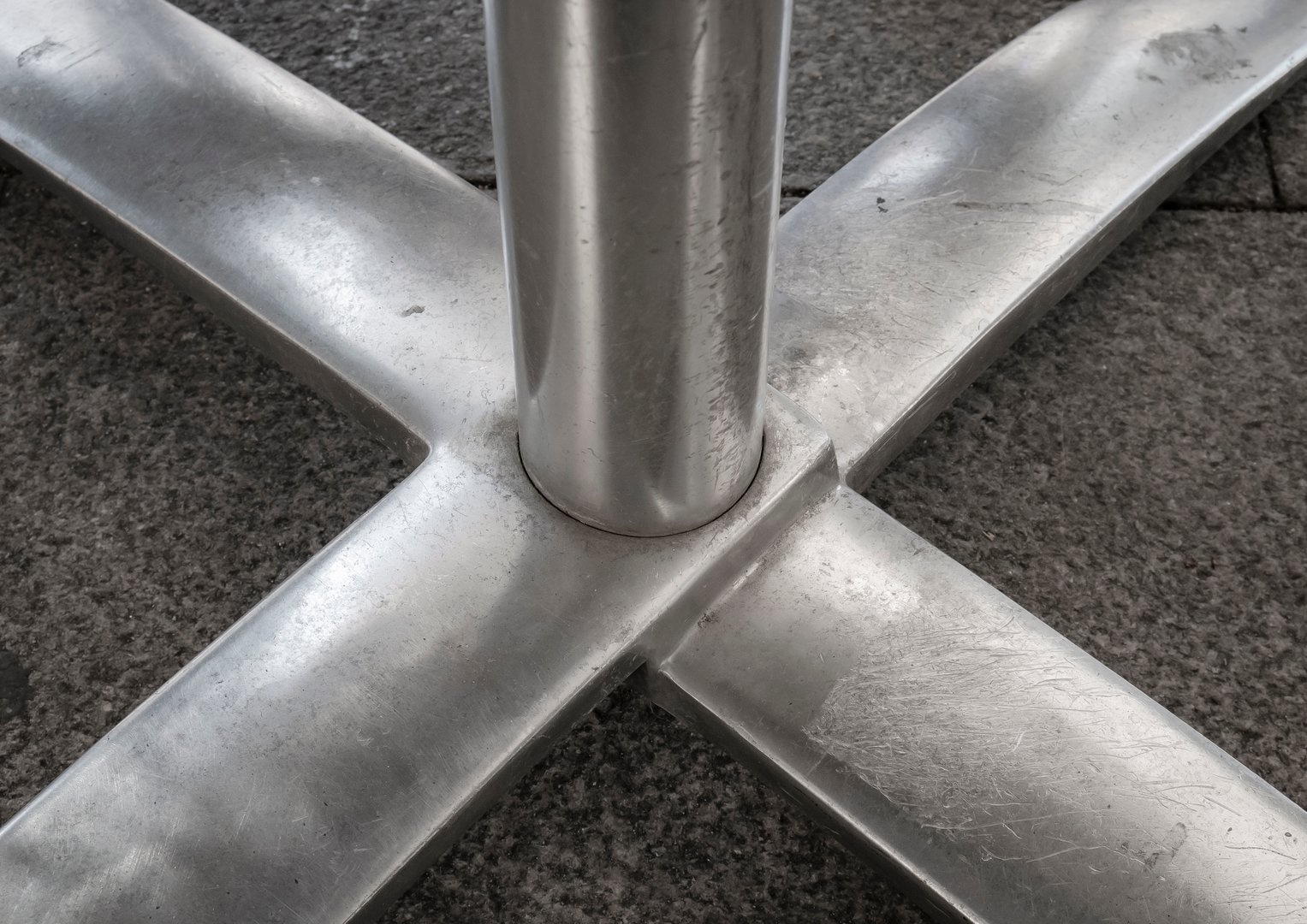 Metal base composed of pieces welded together in the shape of an X and a cylinder that rises towards the table of a cafe terrace located on stone tiles