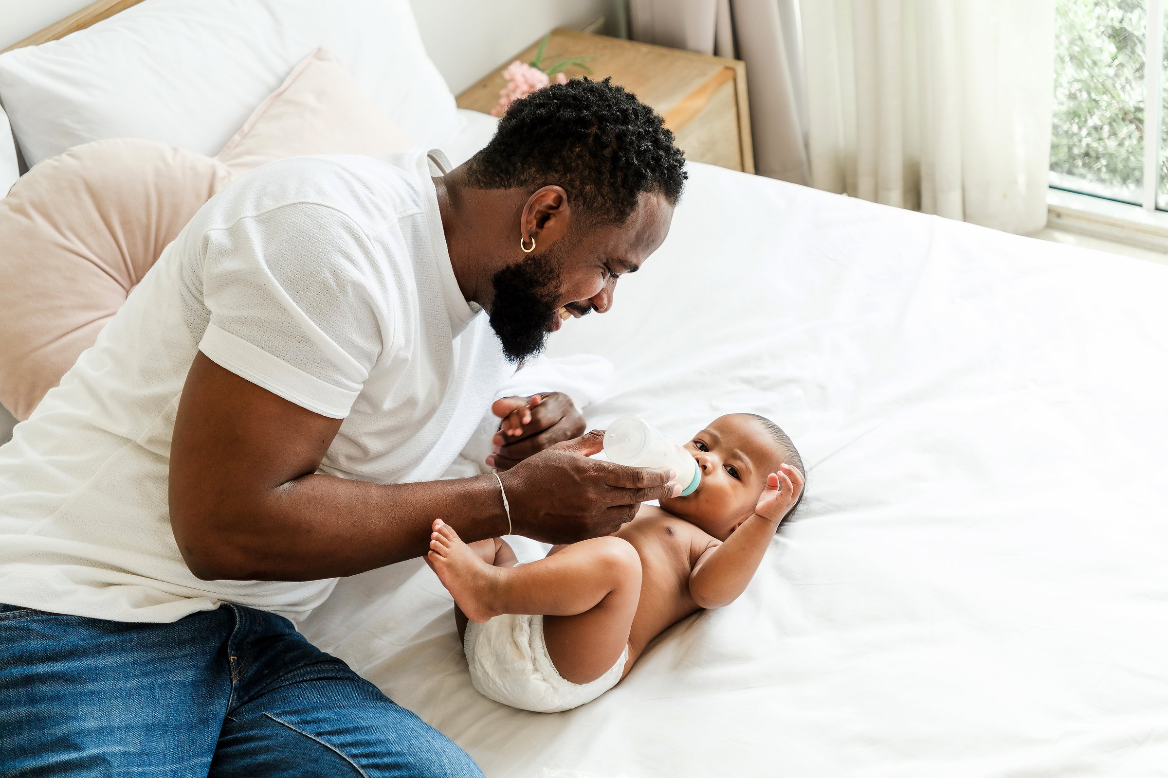 A father joyfully feeds his baby on a cozy bed, highlighting a moment of bonding and care. Happy diversity family and newborn bathing concept.