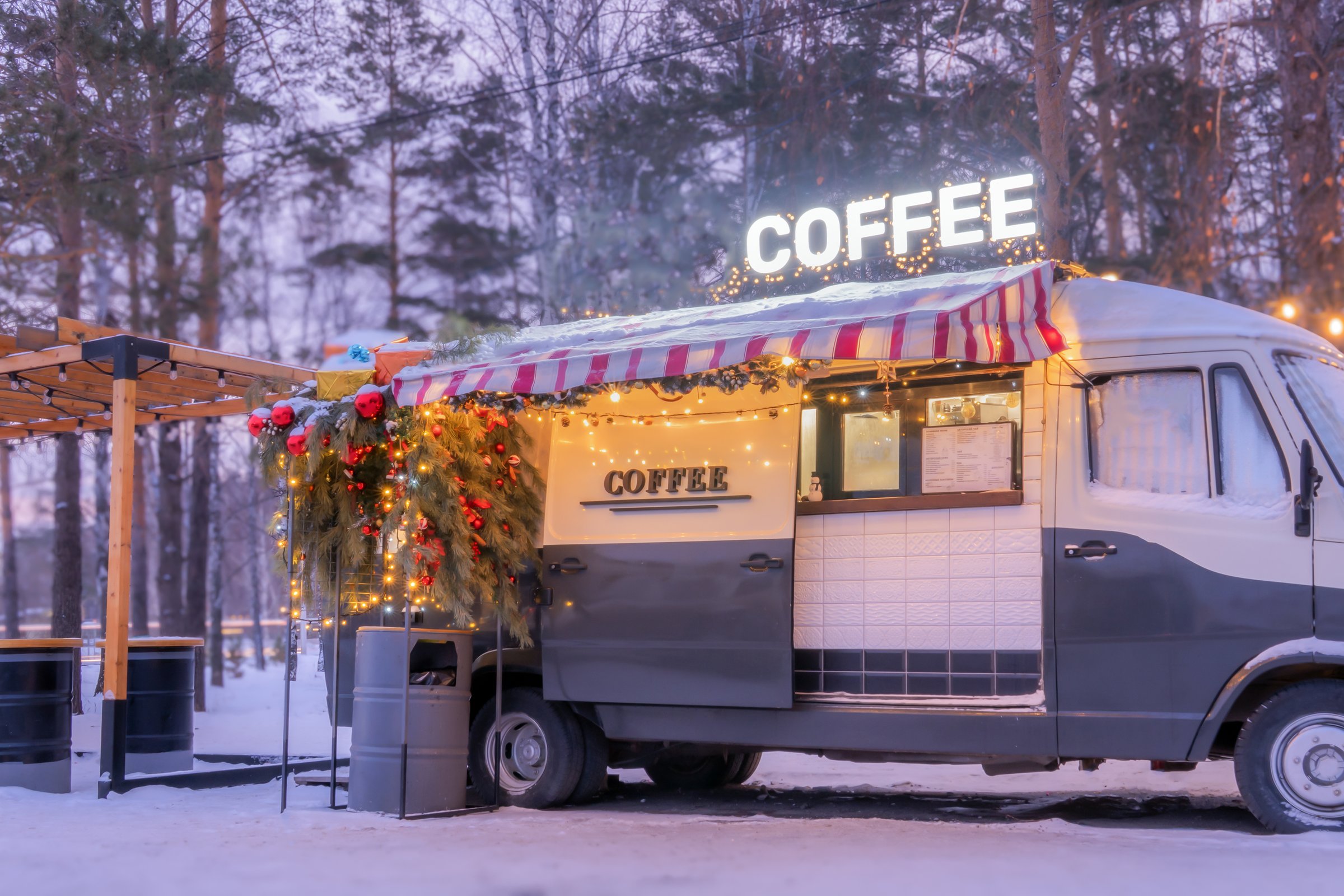 A cozy coffee truck, adorned with festive lights and greenery, stands in a snowy forest. The atmosphere captures the charm of winter as customers enjoy warm beverages