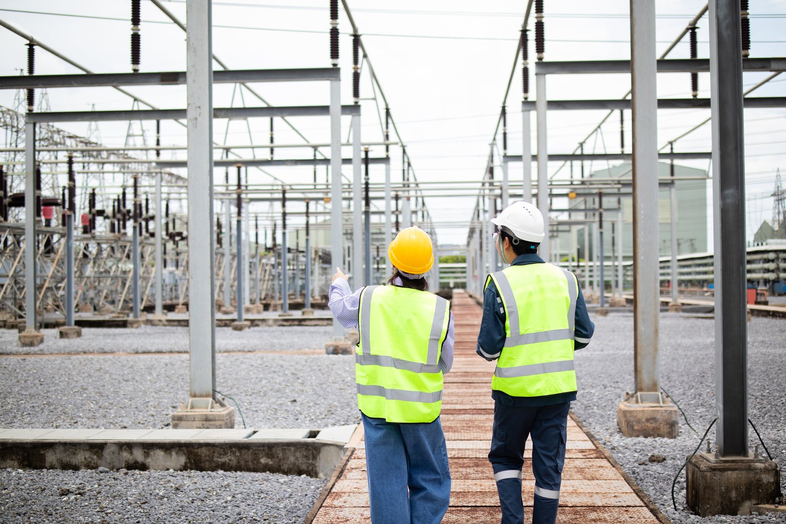 A two engineers are working at high voltage substation wearing safety helmets and reflective vests while inspecting electrical equipment in industrial outdoor setting