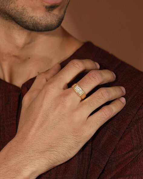 A person wearing a gold ring with a rectangular design, resting hand on a chest, dressed in a burgundy textured top.