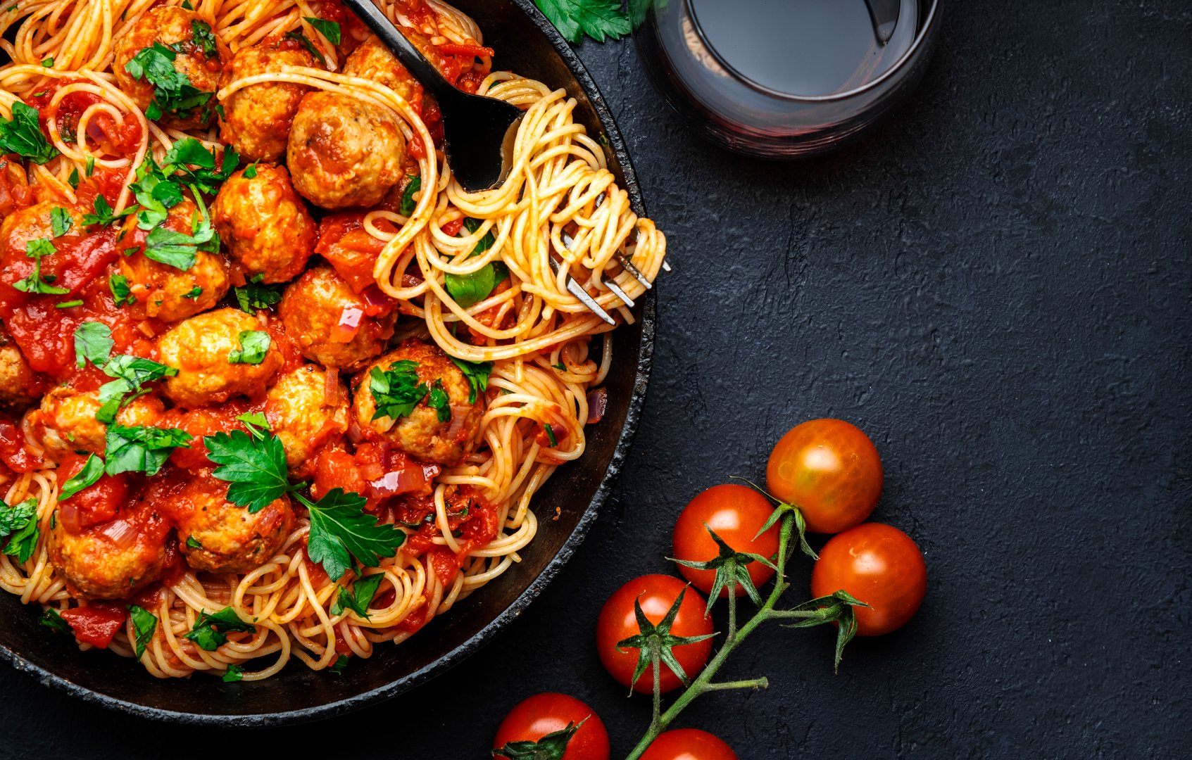 Prepared spaghetti pasta with fried pork meatballs in tomato sauce with parsley in frying pan, black table background, top view