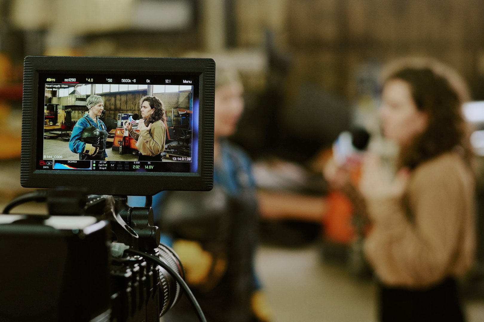 Caucasian middle aged woman and Caucasian young adult woman standing in industrial workshop being filmed by professional camera, monitor displaying both women in conversation