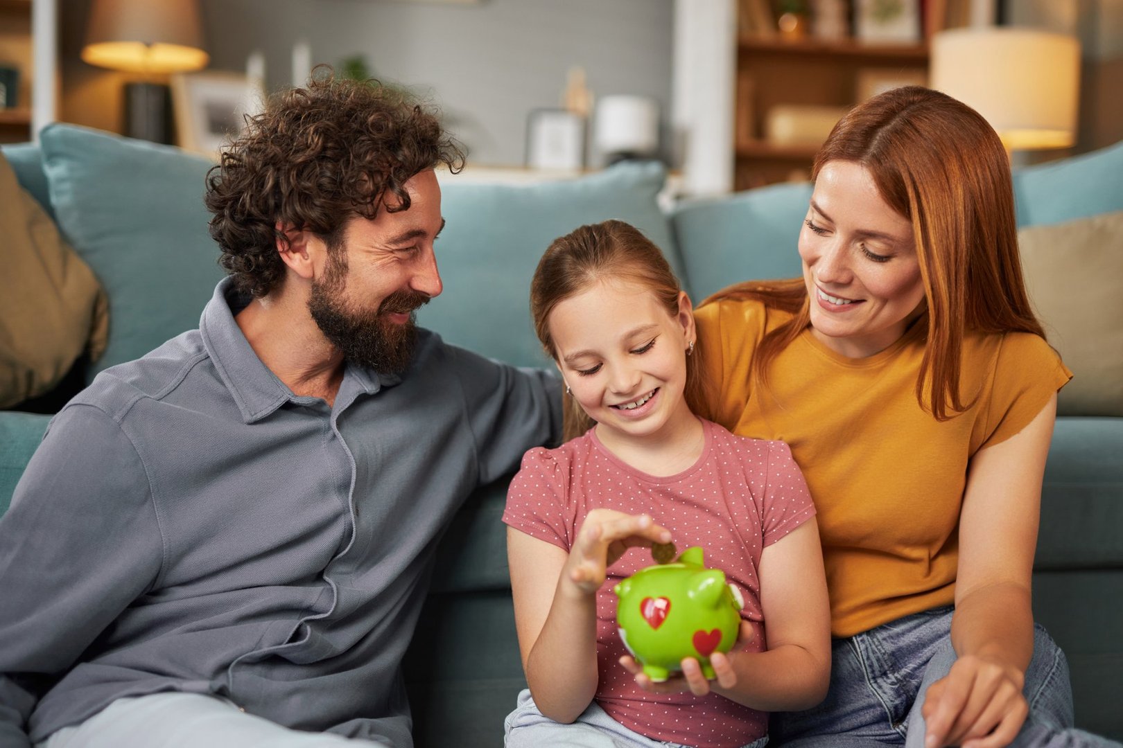 Smiling parents watching their daughter putting a coin into piggy bank, teaching her importance of saving