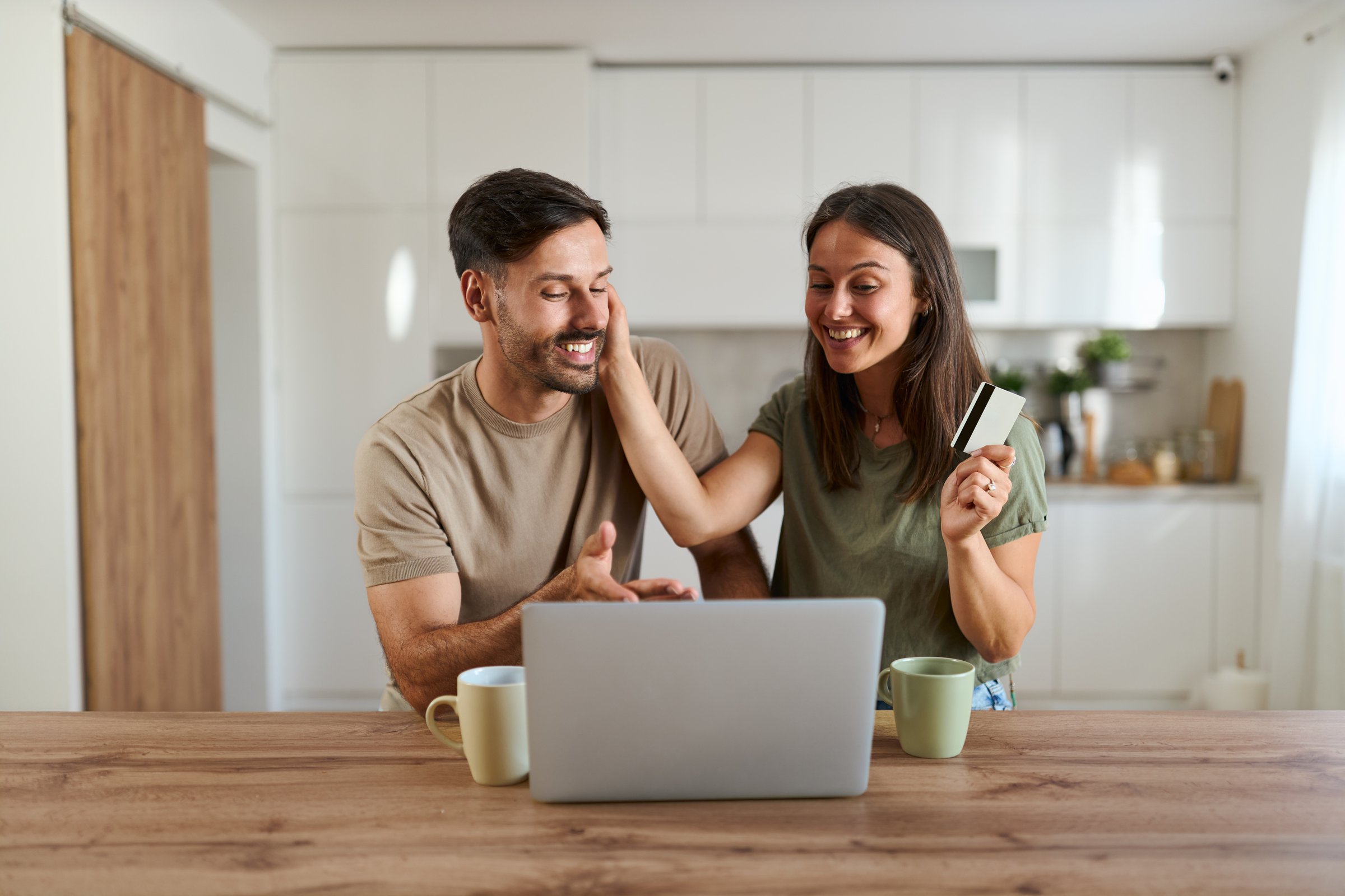 Happy couple using credit card for online shopping over laptop at home and looking at camera.