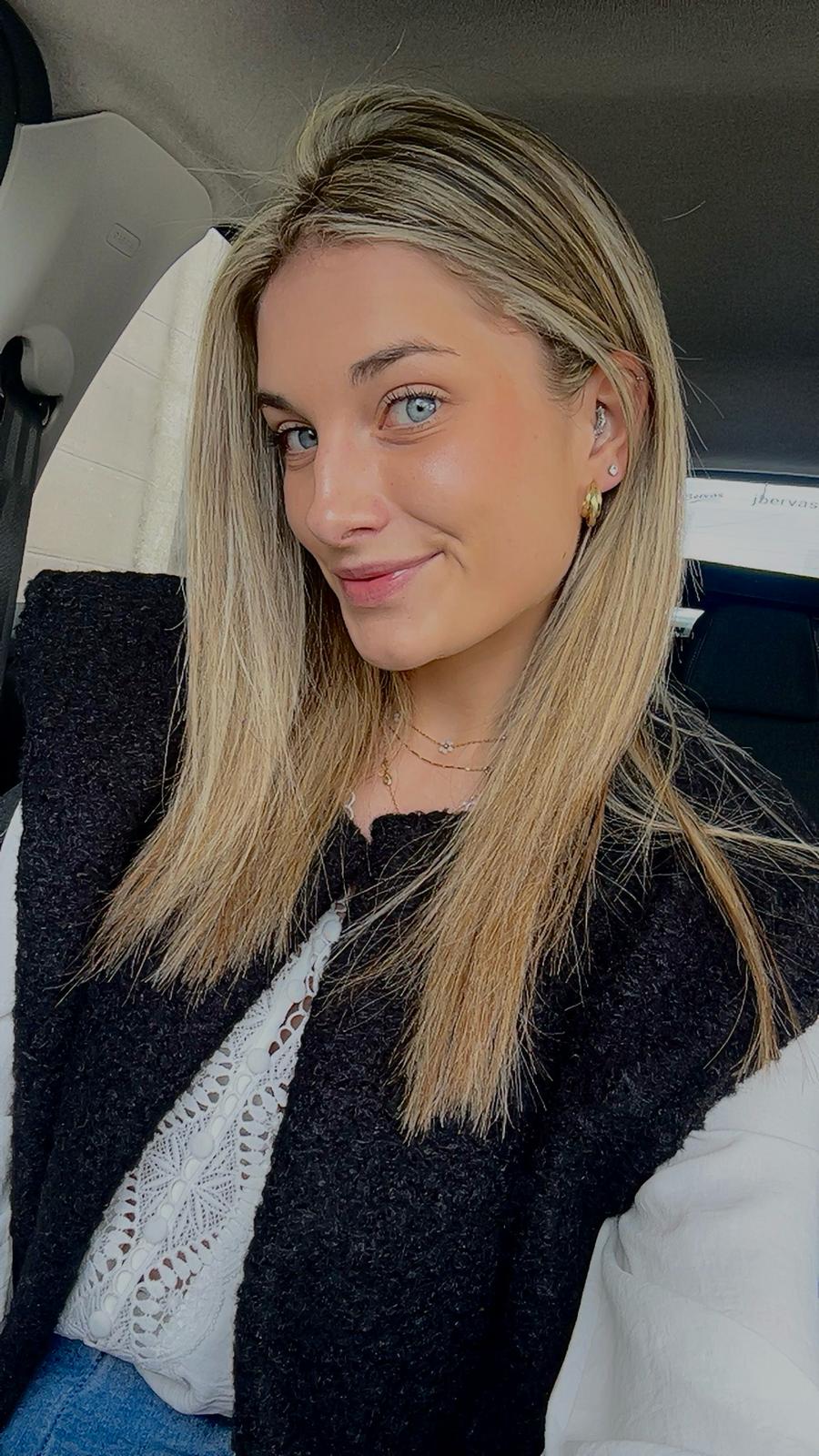 Woman with long blonde hair and blue eyes smiling in a car, wearing a white lace top and black vest.
