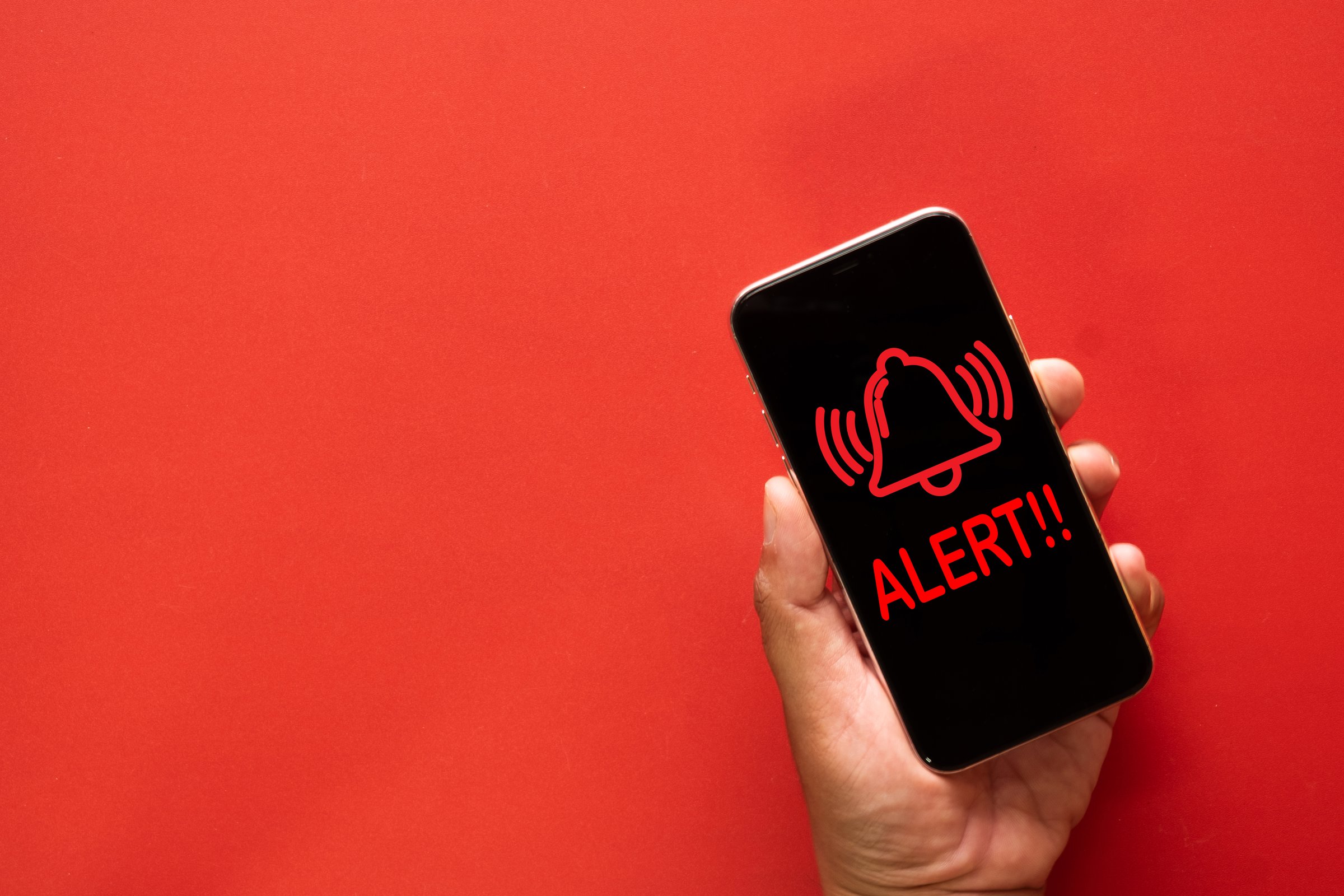 Hand holding mobile phone displaying alert notification, red bell icon with ALERT!! message, phone warning, emergency notification, technology, communication, urgent message concept.