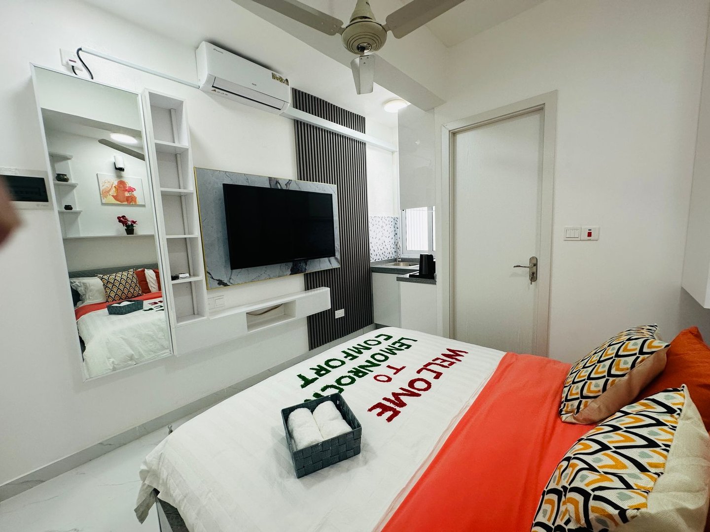 Modern bedroom with a TV, wall-mounted shelves, decorated bed, and small desk area by the window.