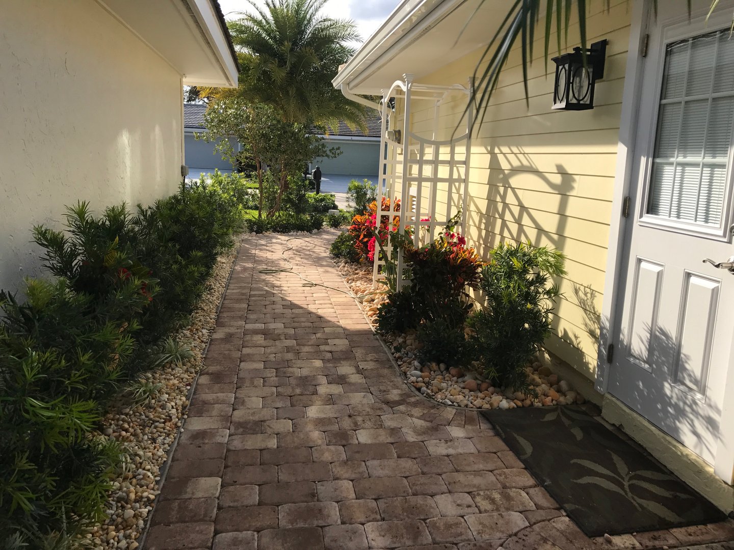 Sunlit brick pathway between yellow house and shrubs, featuring potted plants, stone borders, and a white trellis.