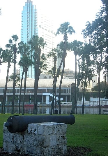A historic cannon on a stone base in a park with palm trees, with a tall modern building in the background near a body of water.