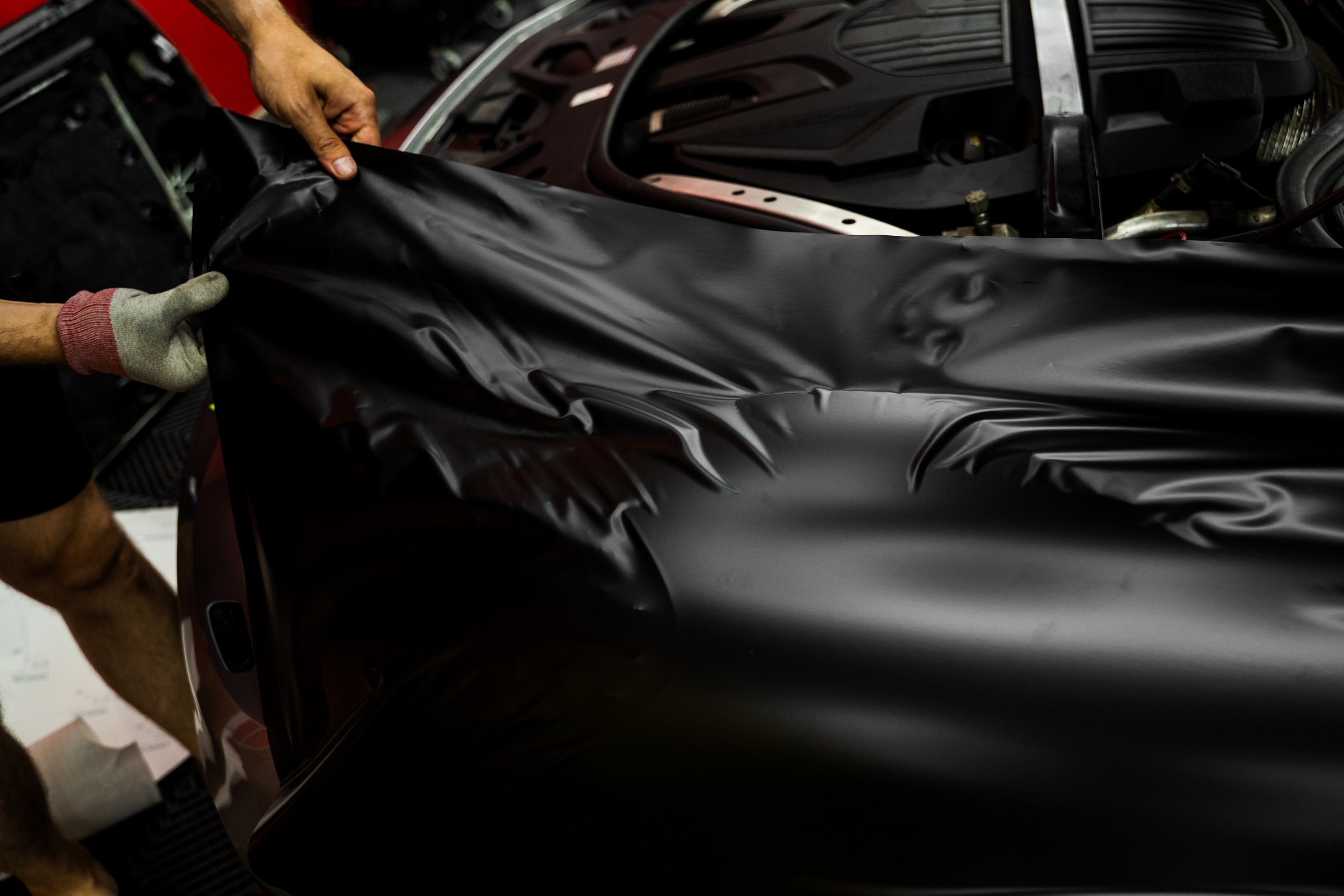 Careful application and tensioning of black vinyl avoids surface damage or bubbling. Car wrapping process using black matte vinyl stretched into place. Matte black vinyl is applied across car hood