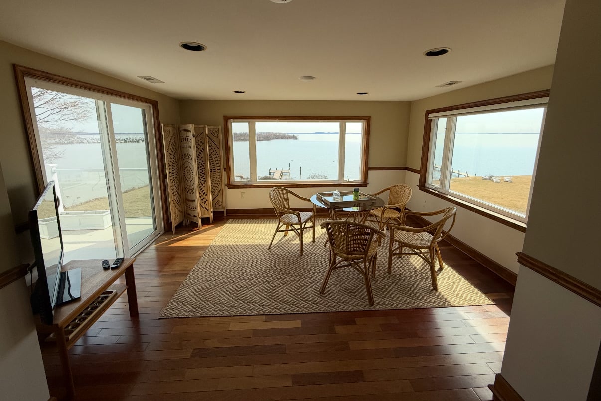 Room with wicker chairs and table, large windows with water views, wooden floor, and a television on a stand.