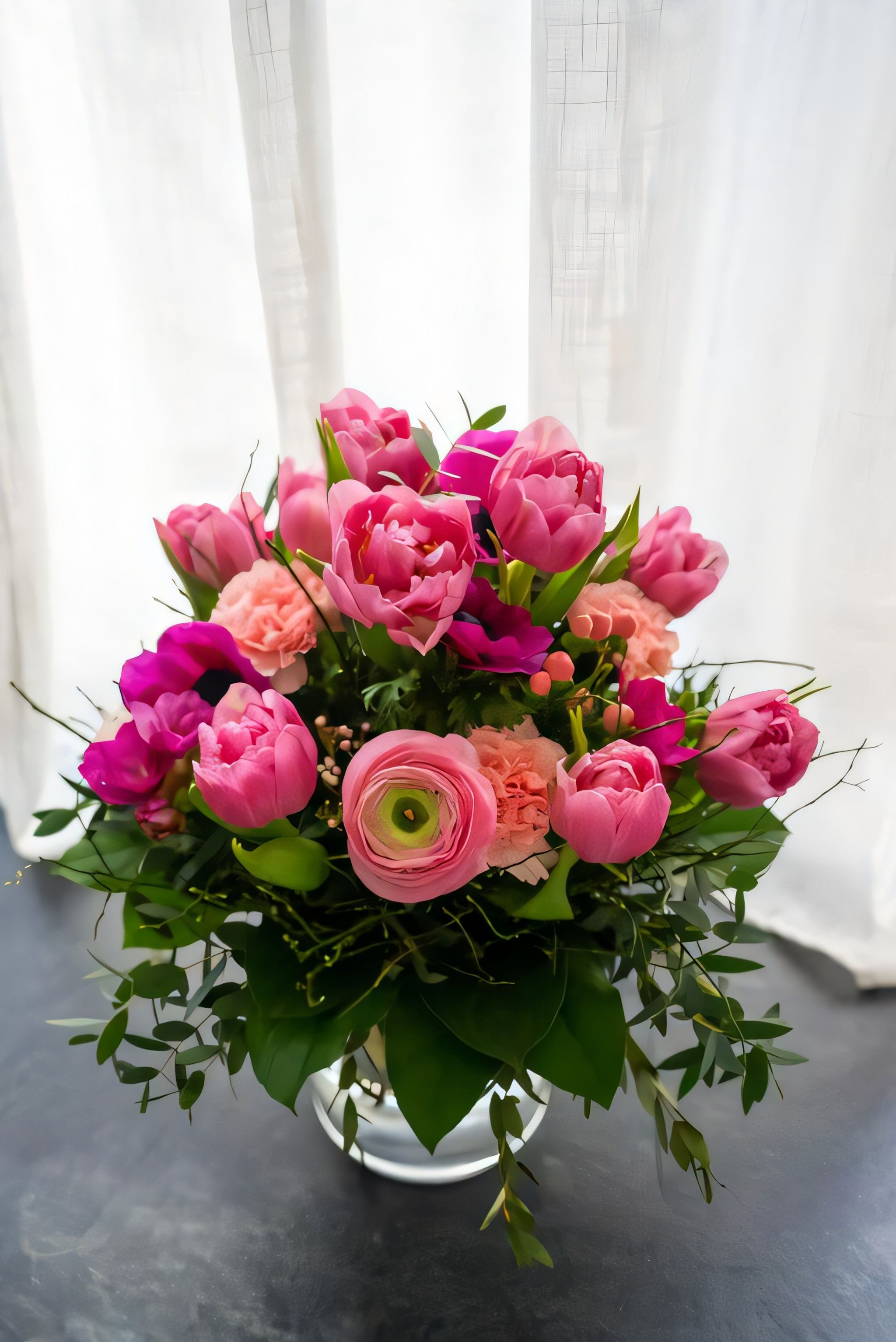 A vibrant bouquet of pink and peach flowers with lush green leaves set against a soft curtain backdrop.