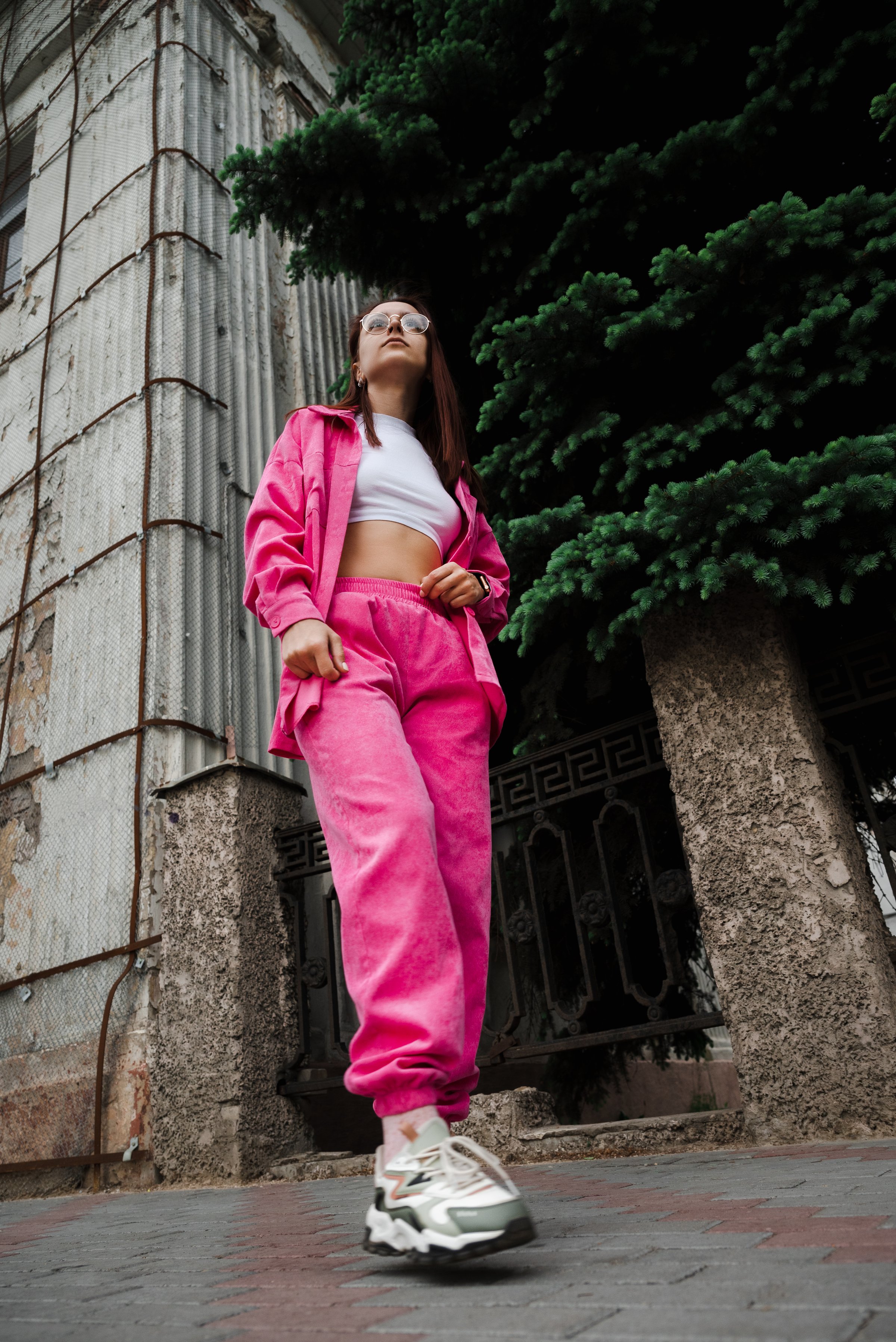 Woman in a pink tracksuit and glasses stands confidently in front of a restoration building, showcasing a trendy, urban style.