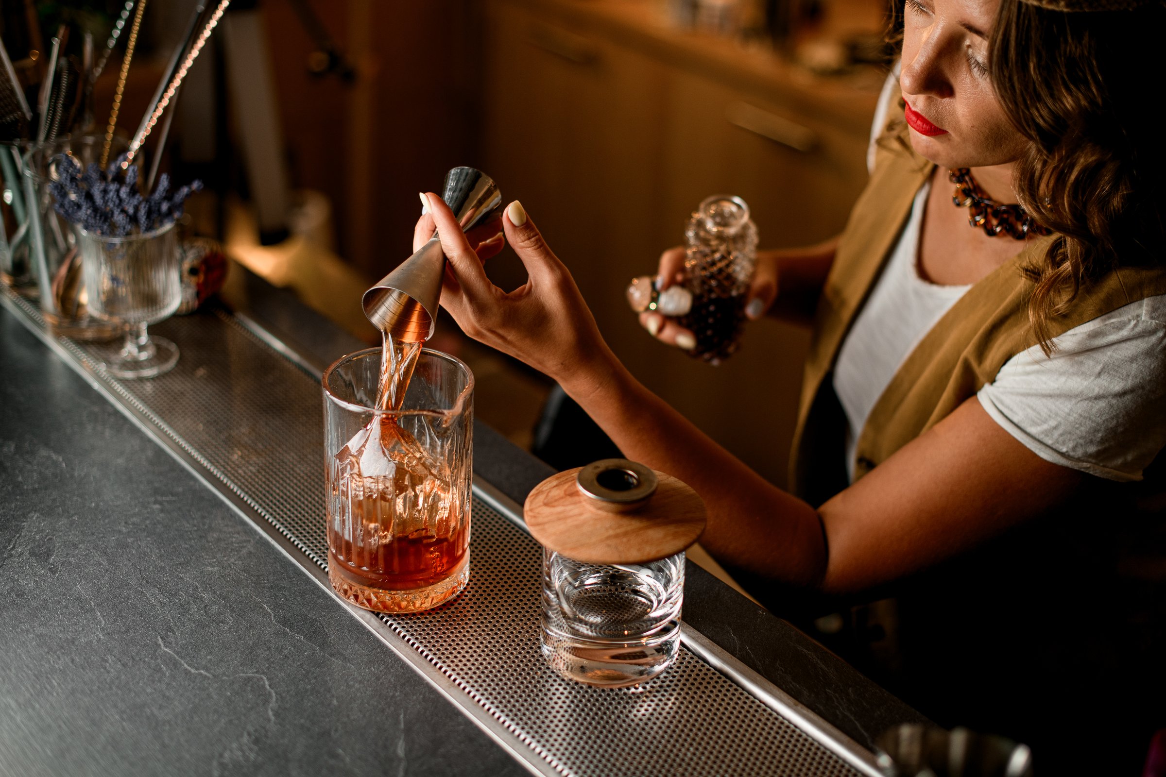 Top view of bar counter with mixing glass in which female bartender pours brown drink from jigger