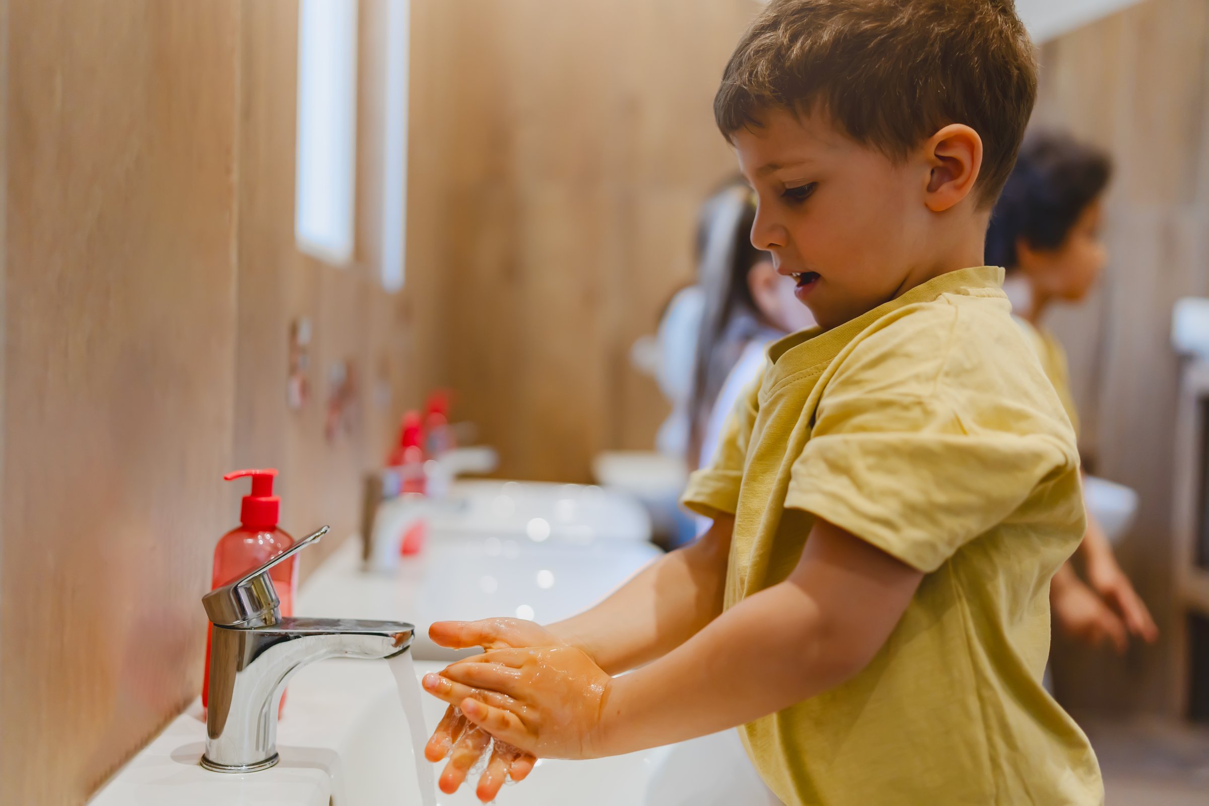 A preschool boy washes his hands at the sink, practicing hygiene with other children in a kindergarten bathroom.