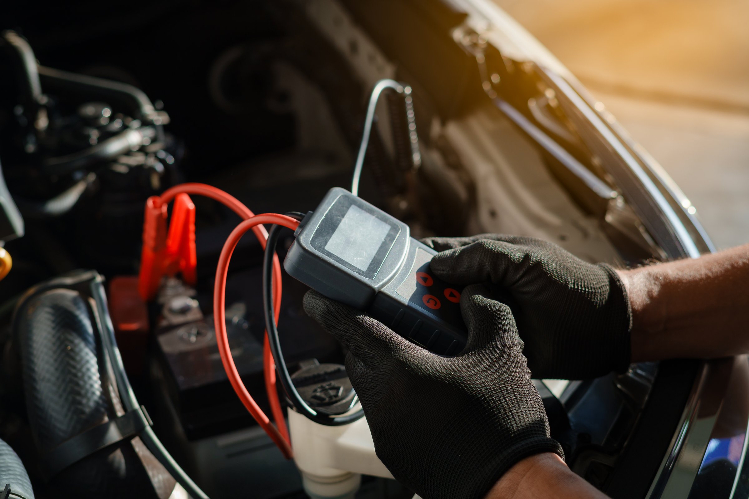 Professional car mechanic using a digital battery tester to inspect vehicle performance during routine maintenance. Automotive technician wearing gloves performing diagnostics under the hood in a repair shop.