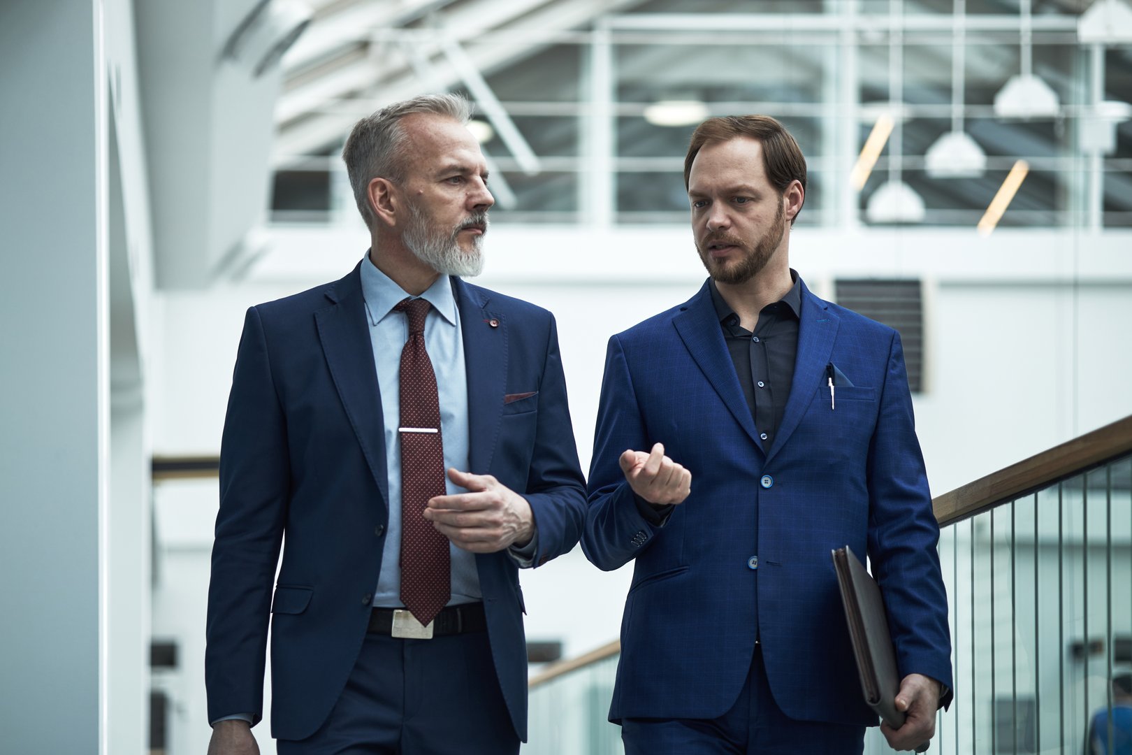 Two businessmen walking and engaging in conversation inside a modern office building with glass elements and open interiors. Both wearing formal blue suits and holding documents