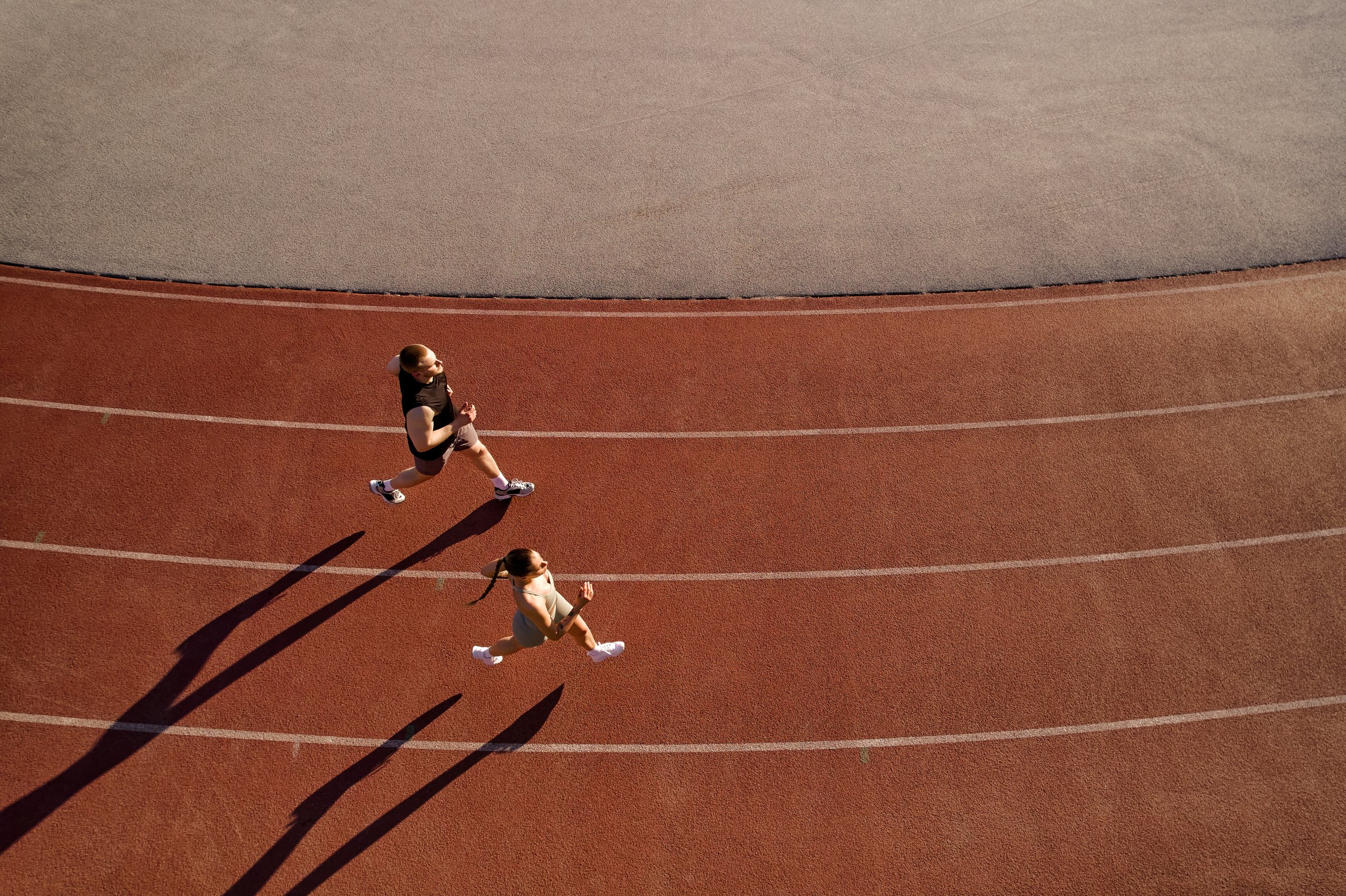 Drone shot of a man and woman running on a curved red track, casting long shadows in evening light. Concept of fitness, teamwork, and determination.