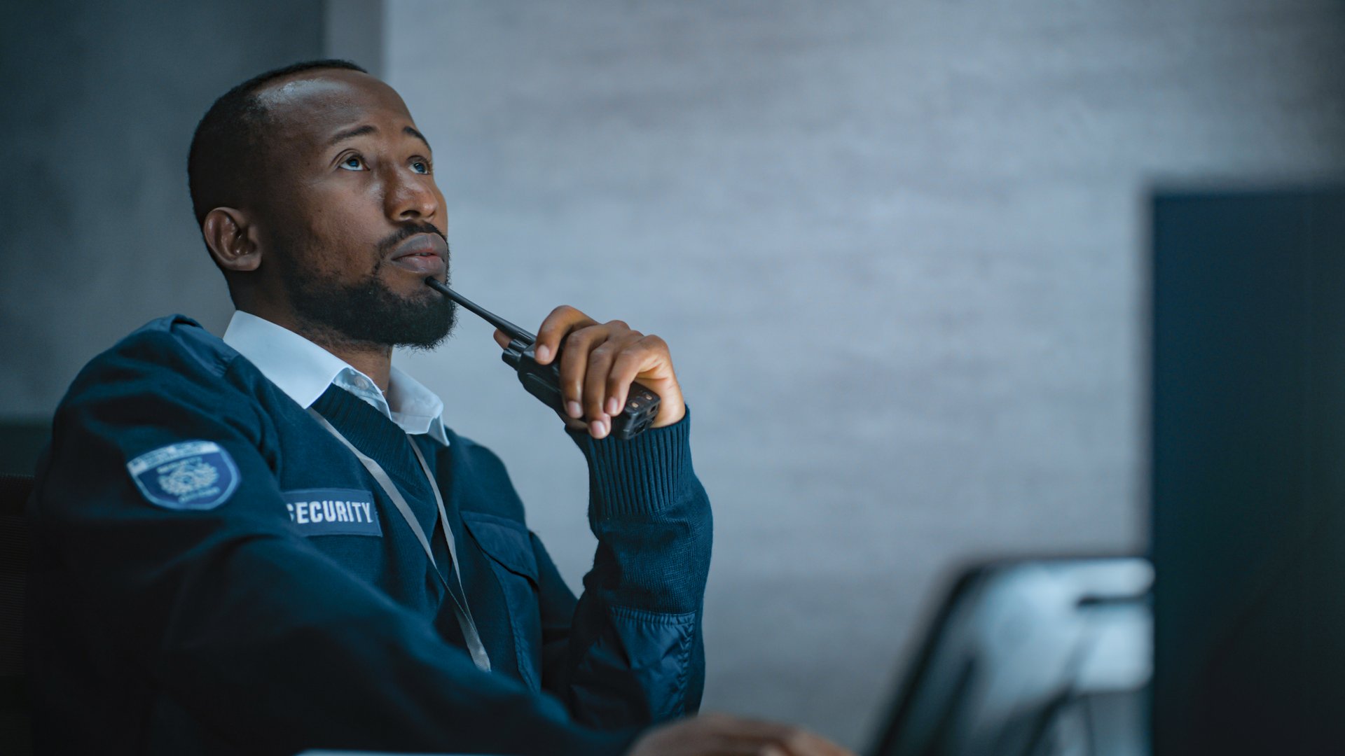 African American security officer or dispatch in uniform uses tablet computer and walkie talkie, looks at screens working in monitoring center during night shift. Surveillance and CCTV system concept.
