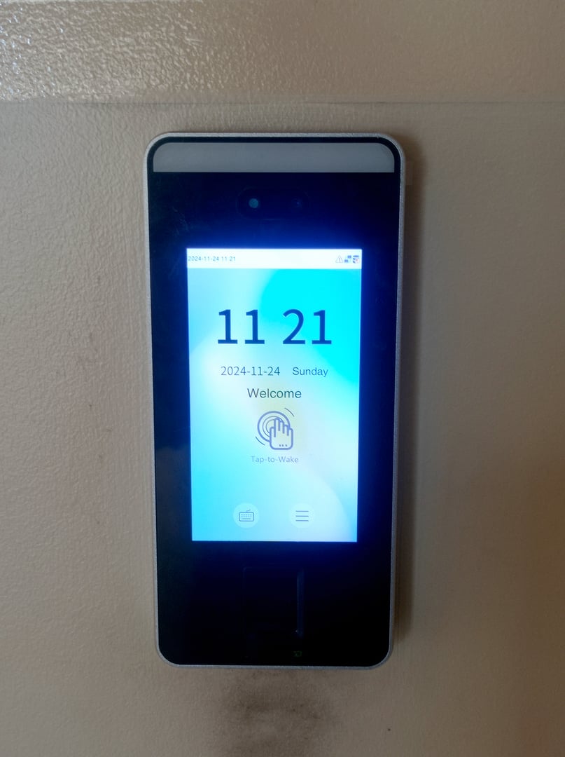 A close-up view of a sleek, black digital access control device mounted on a wall, displaying the time 11:21 and a welcome message in bright blue light.