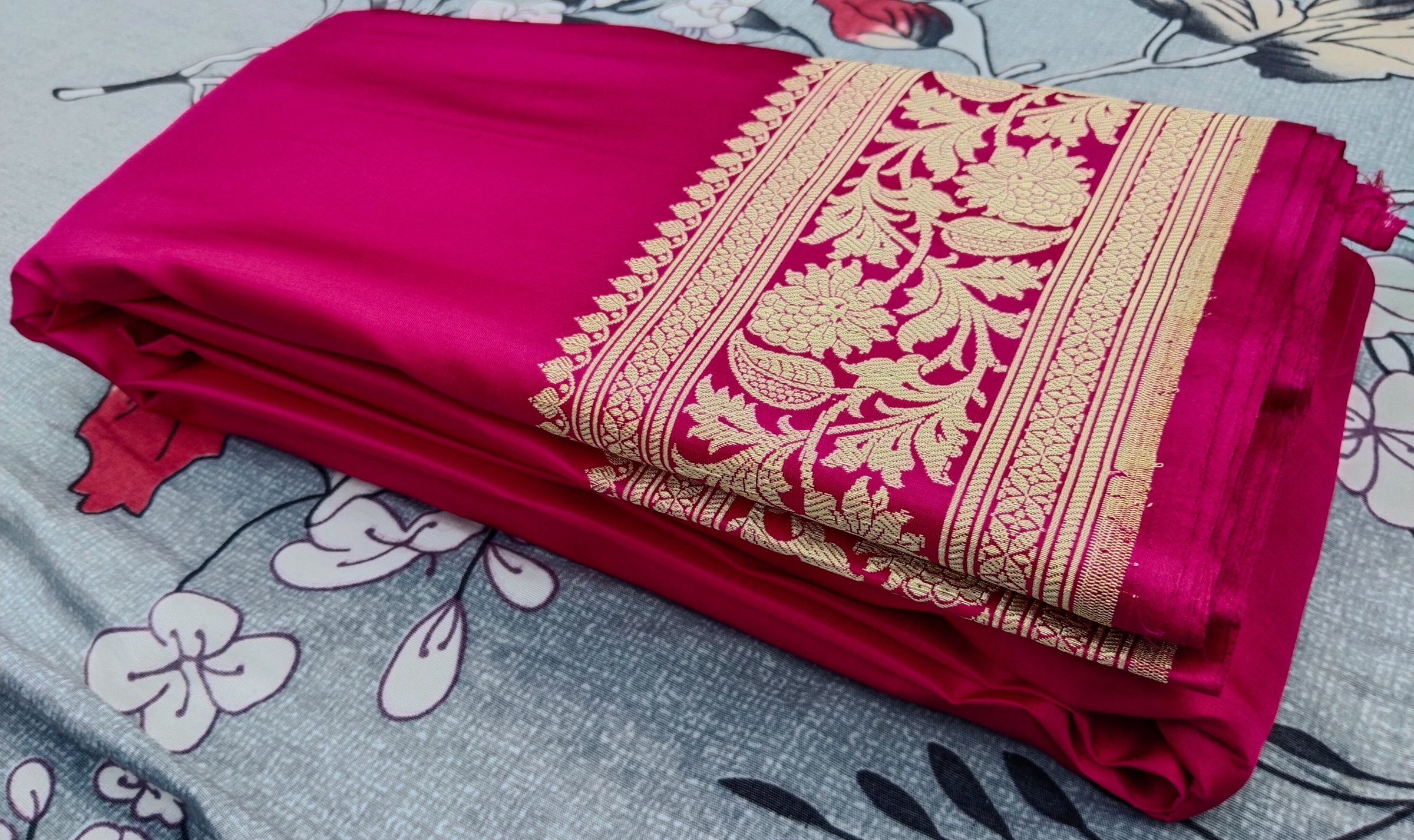 Folded dark pink Banarasi silk saree with intricate golden zari floral border placed on a floral bedsheet, showcasing rich traditional Indian textile and ethnic wedding fashion elegance