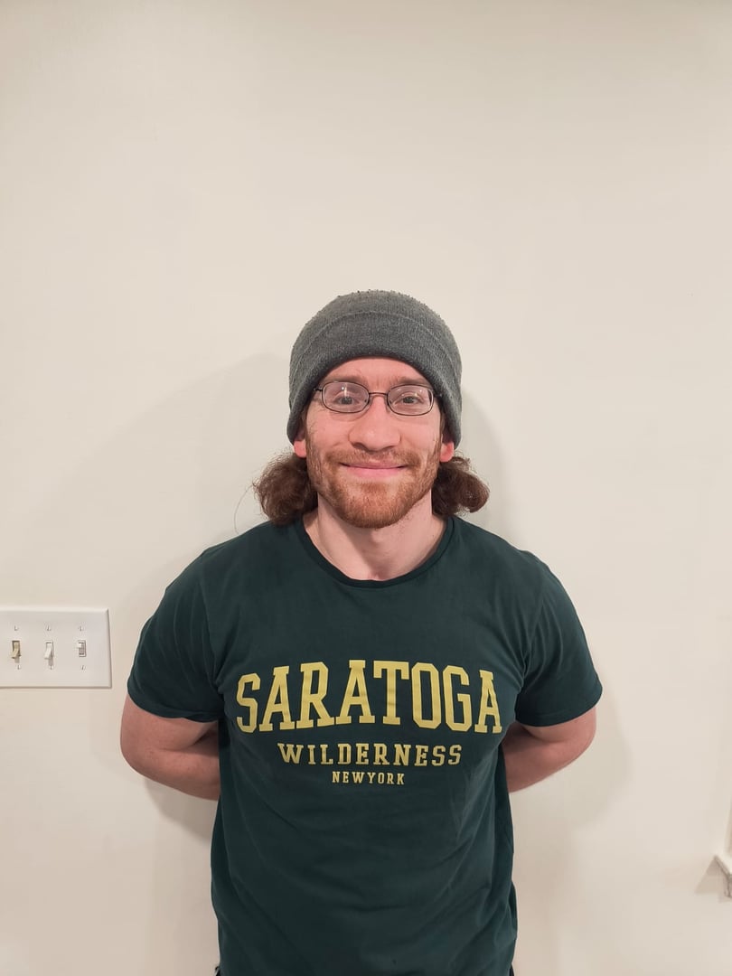 Person with long hair, glasses, and a beanie, wearing a green Saratoga Wilderness New York T-shirt, standing against a wall.