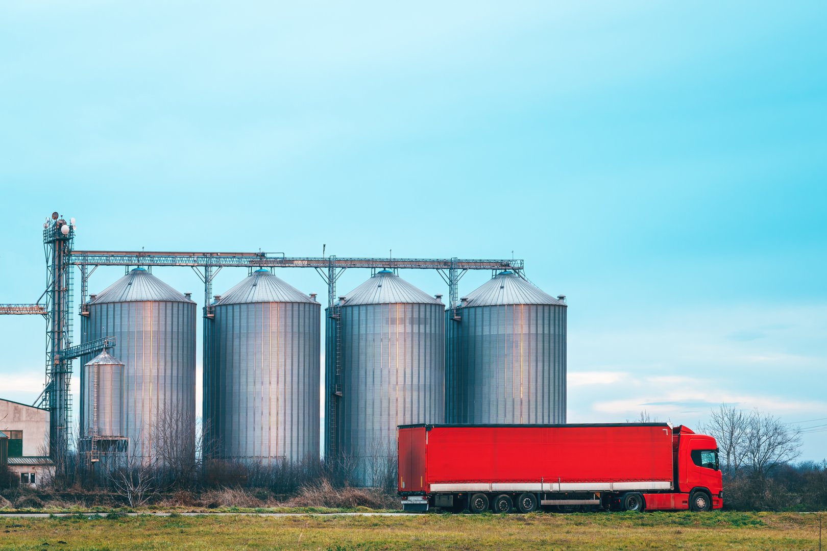 Semi-truck with red trailer in front of agricultural grain processing plant silos, selective focus