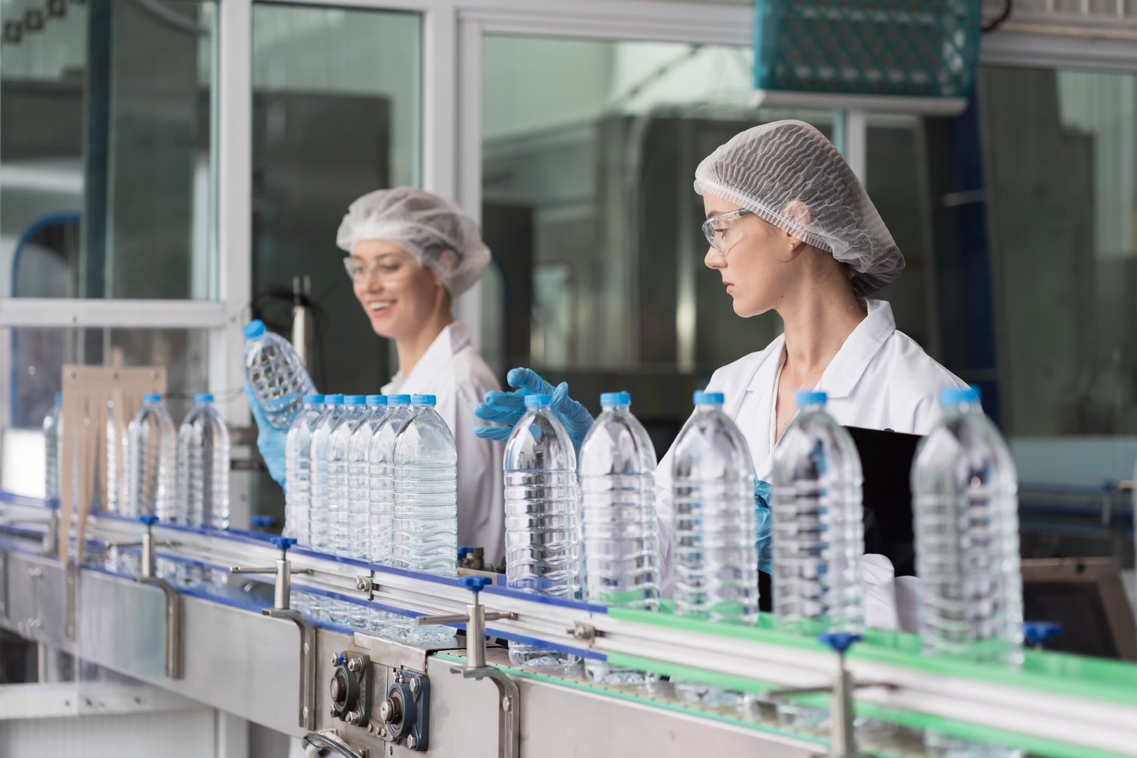 Two female worker working inspecting quality clean purified drinking plastic bottled water inside automated conveyor belt production line in drinking water factory