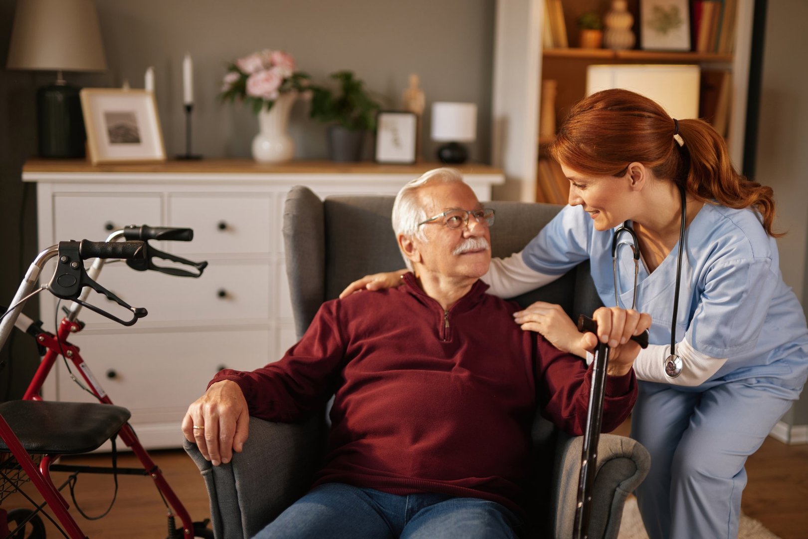 Home healthcare nurse assisting a senior man holding a walking cane, providing support and care in his living room