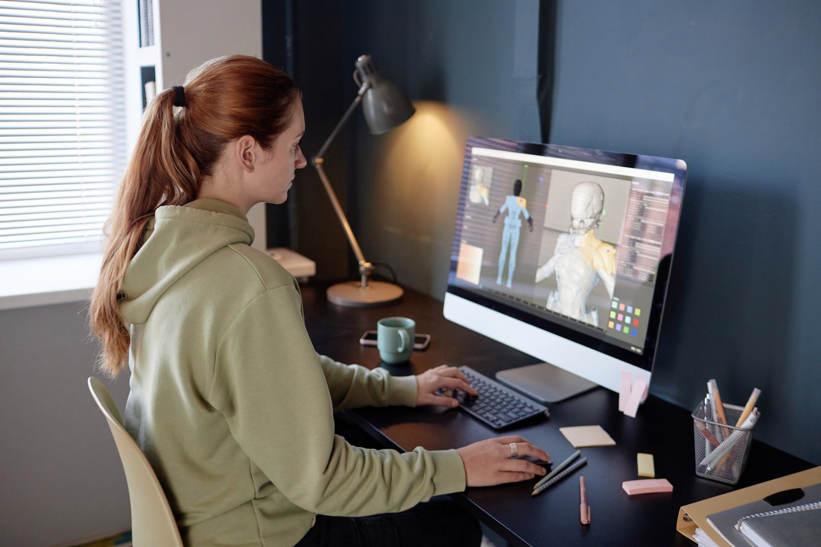 Person sitting at desk creating 3D animation using digital tablet and desktop computer in modern workplace. Lamp and stationery items visible on desk while window light illuminates space