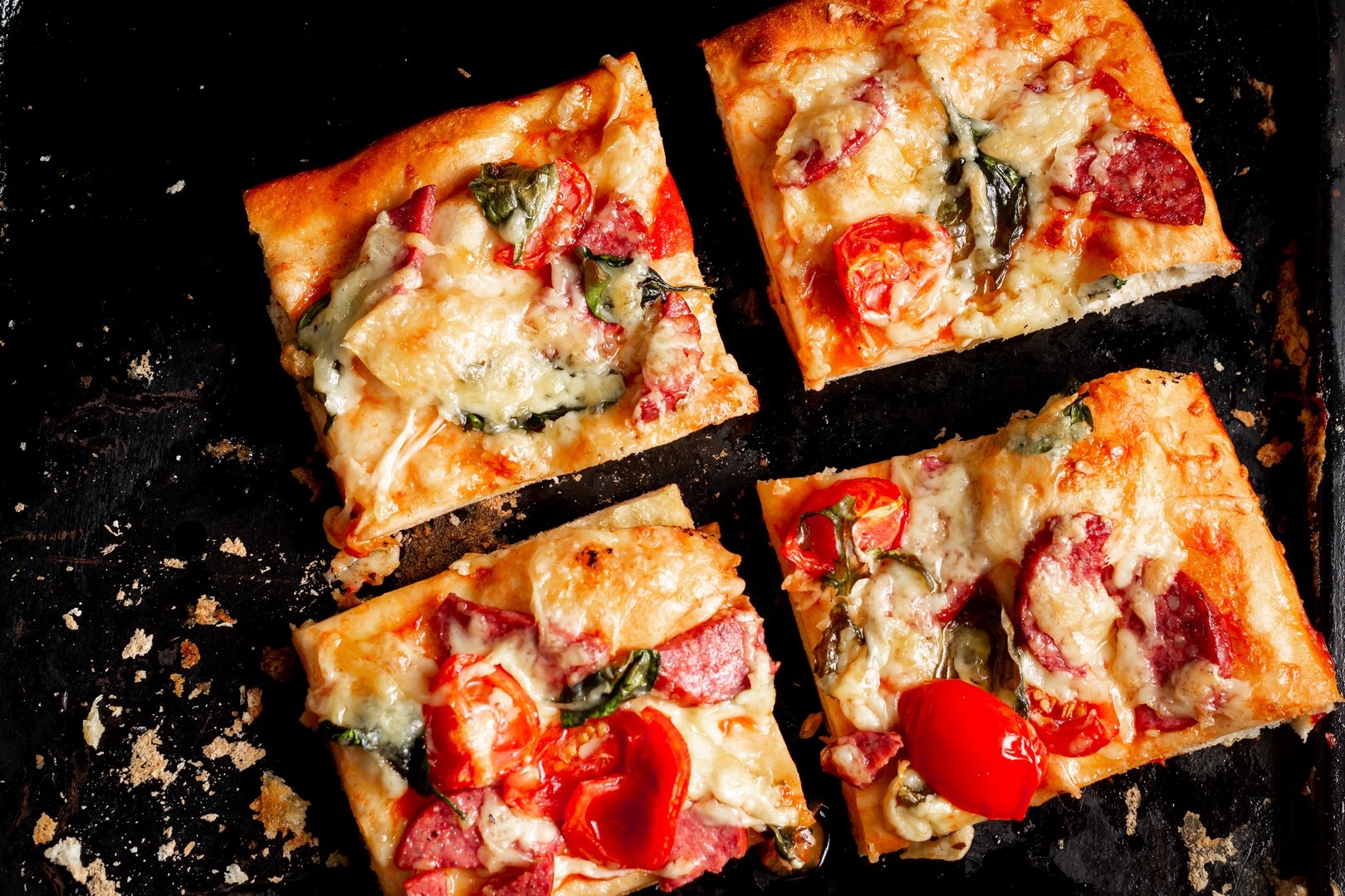 Sliced square pieces of pizza, pie, with tomatoes, sausage, cheese cooked in the oven on a black baking sheet.