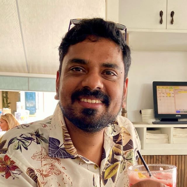 Person smiling, sitting indoors, wearing a floral shirt, holding a drink with a straw. Background features a computer screen.