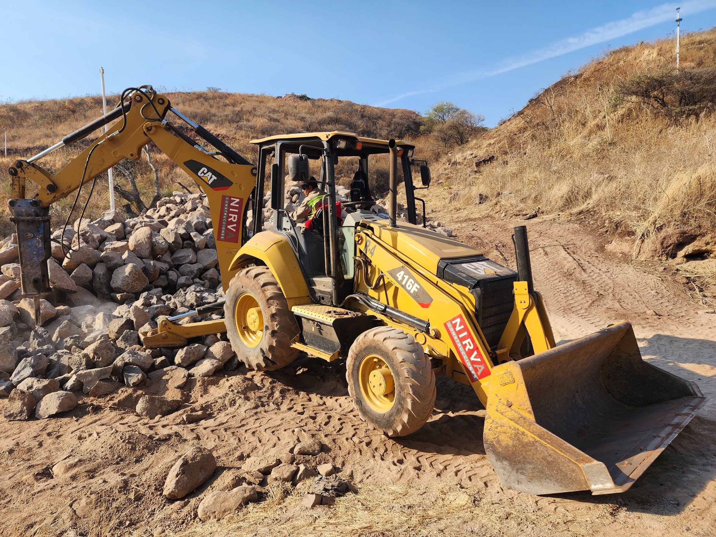 A yellow backhoe loader moving rocks on a dirt path in a hilly, dry landscape with clear blue skies.