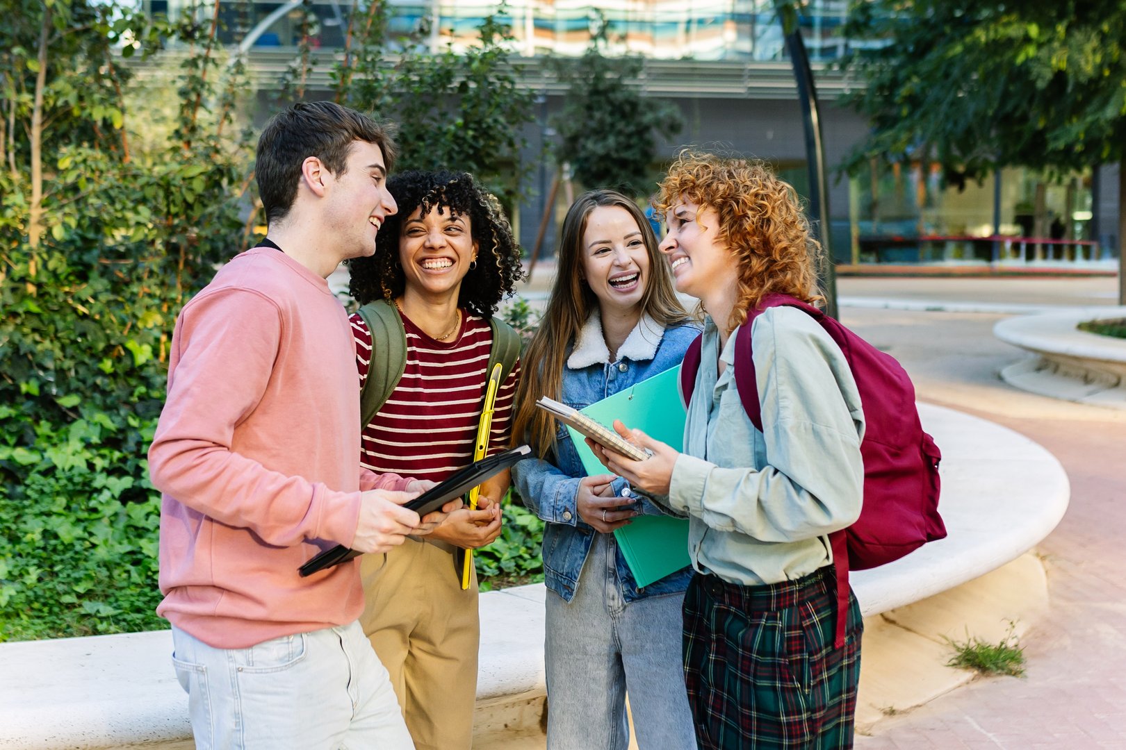 Four diverse young college students laughing together outside building university campus, enjoying their time and sharing a moment of happiness after classes. Education lifestyle and youth concept