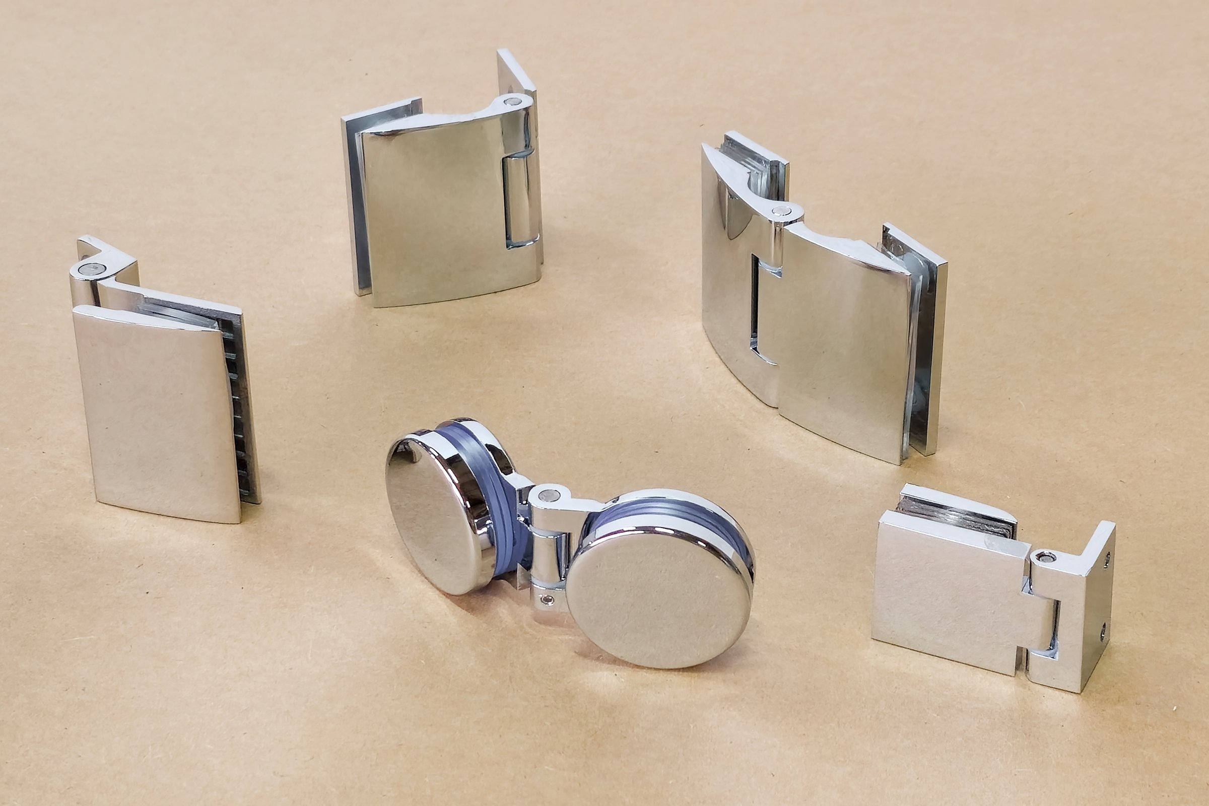 A variety of chrome glass hinges are laid out on a brown table. Necessary accessories for fixing glass doors