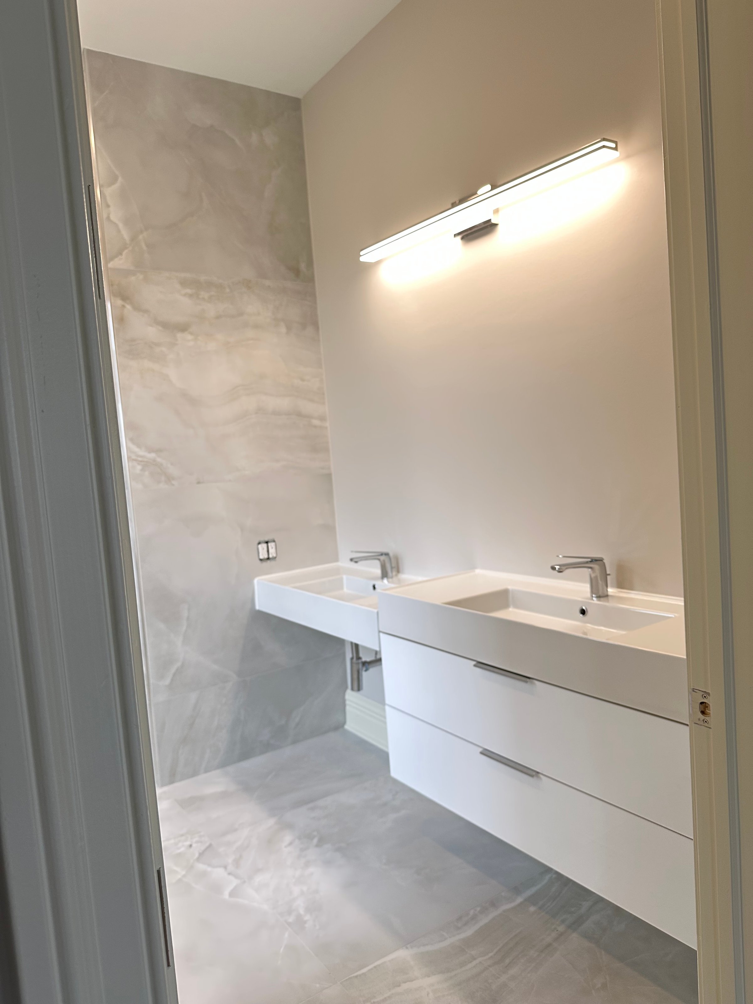 Modern bathroom with dual white sinks, large mirrored wall, and bright overhead lighting on a light gray tiled floor.