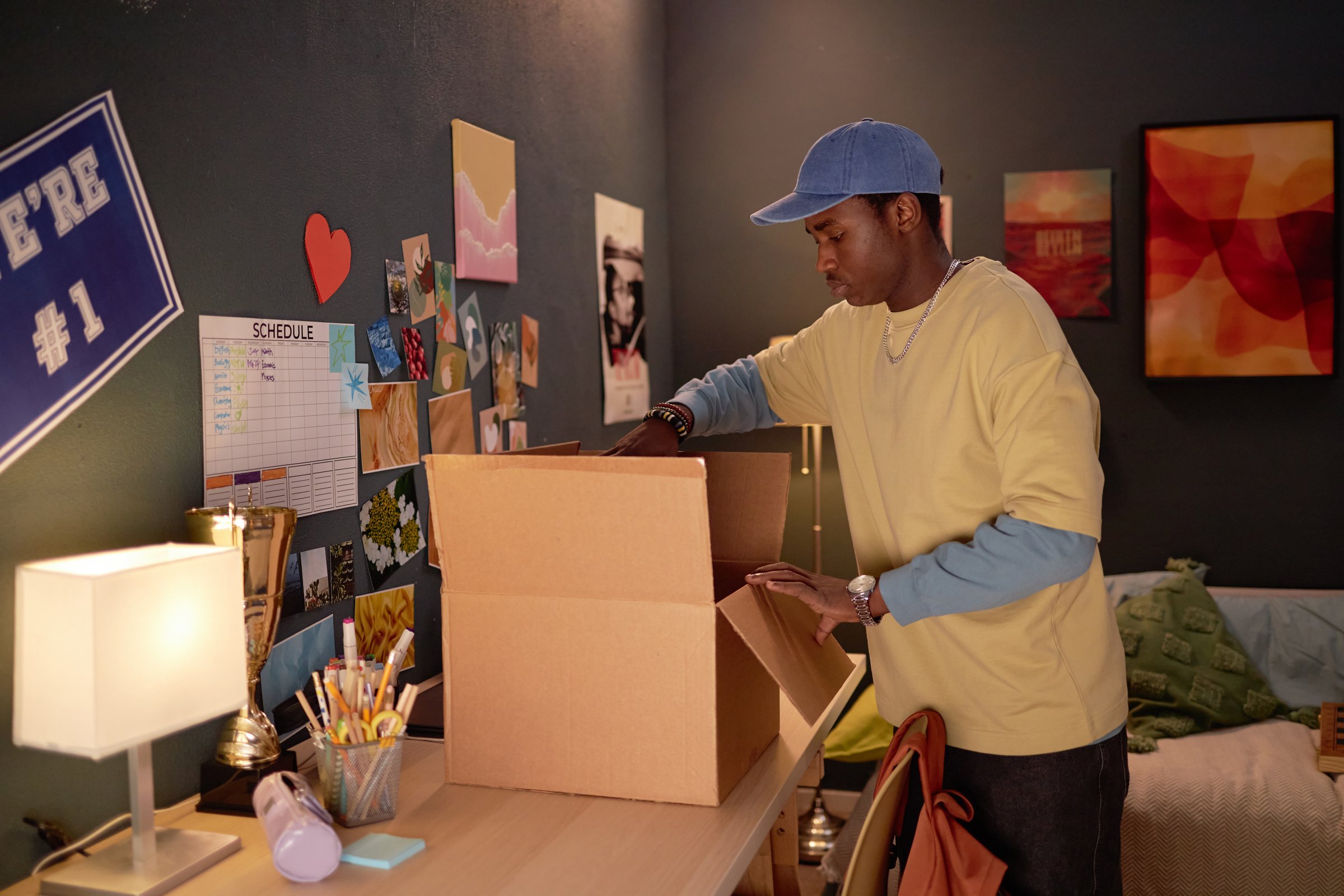Man packing a cardboard box in his dorm room with various items on desk and wall. Neatly arranged schedule board and motivational poster add personal touch