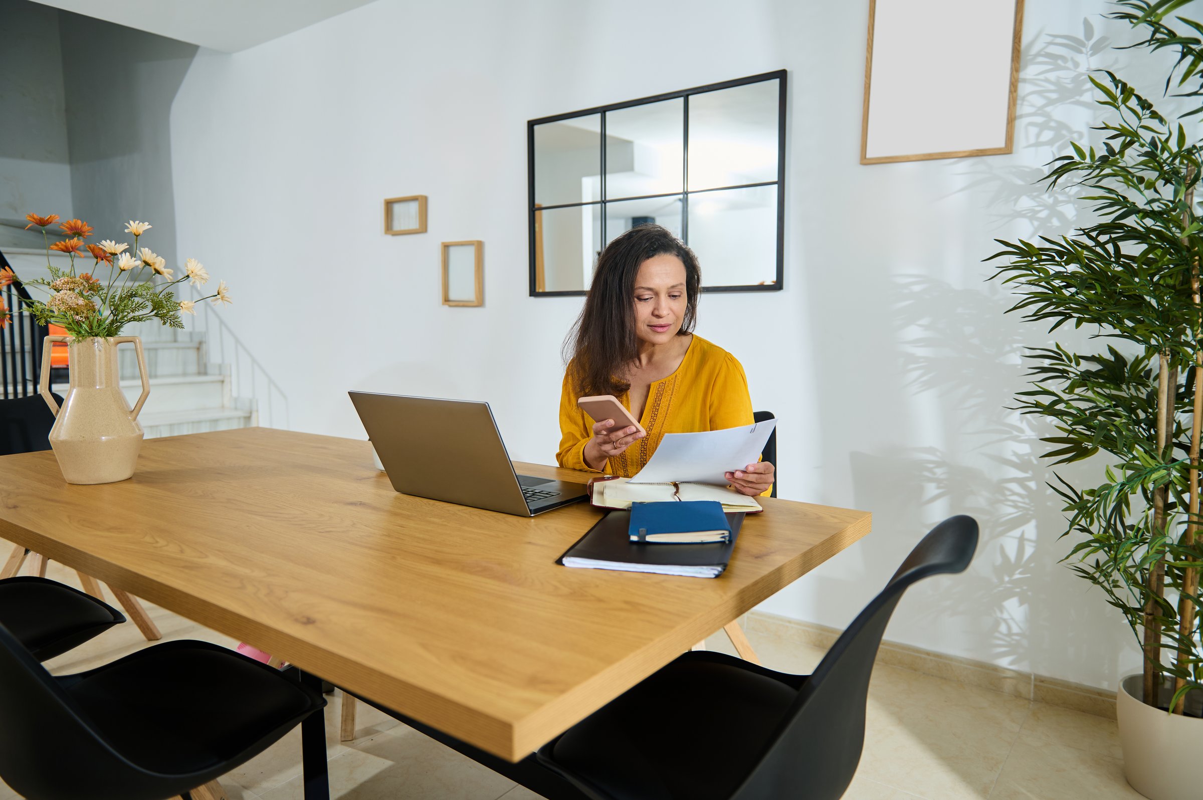 Confident businesswoman working remotely in a stylish home office setting, reviewing documents, managing tasks using laptop and phone, promoting professionalism and comfort in a productive environment