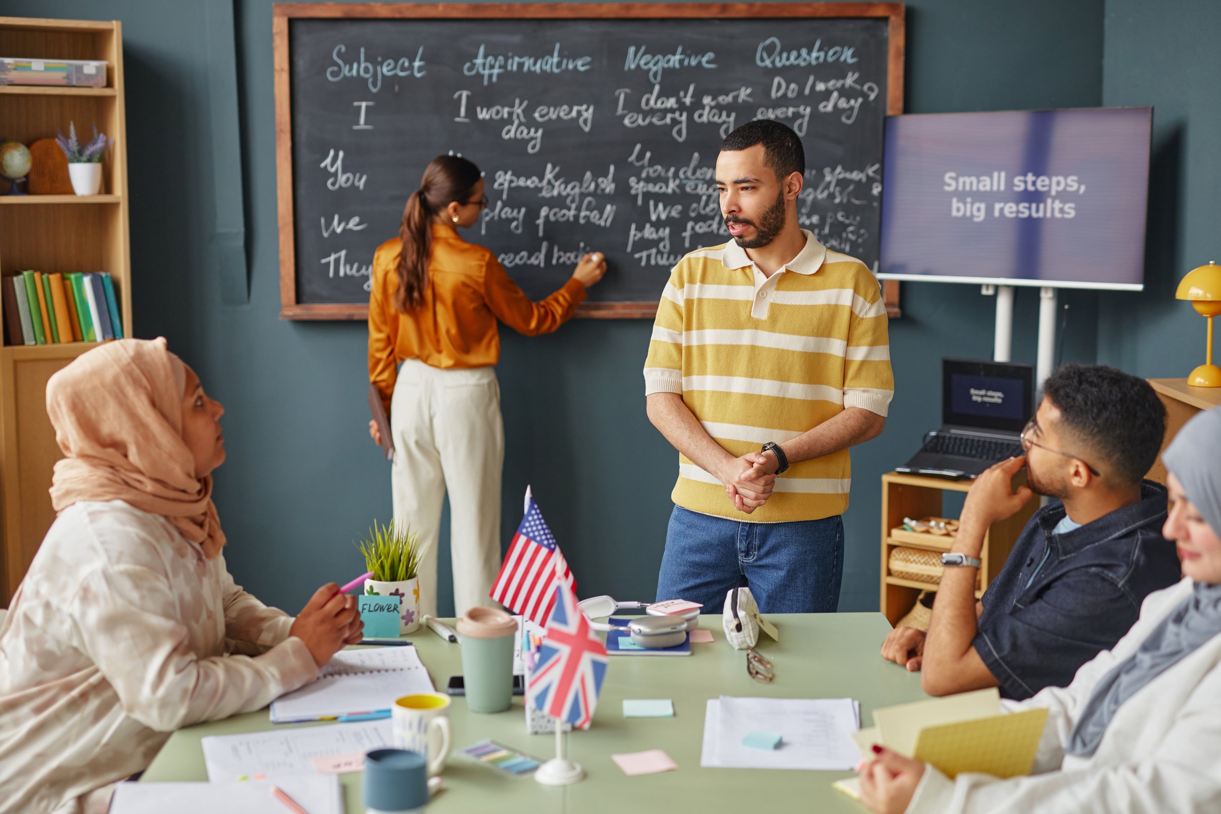 Diverse group of adult men and women attending language school lesson, Middle Eastern man interacting with classmates while female teacher writing on chalkboard, British and American flags on table