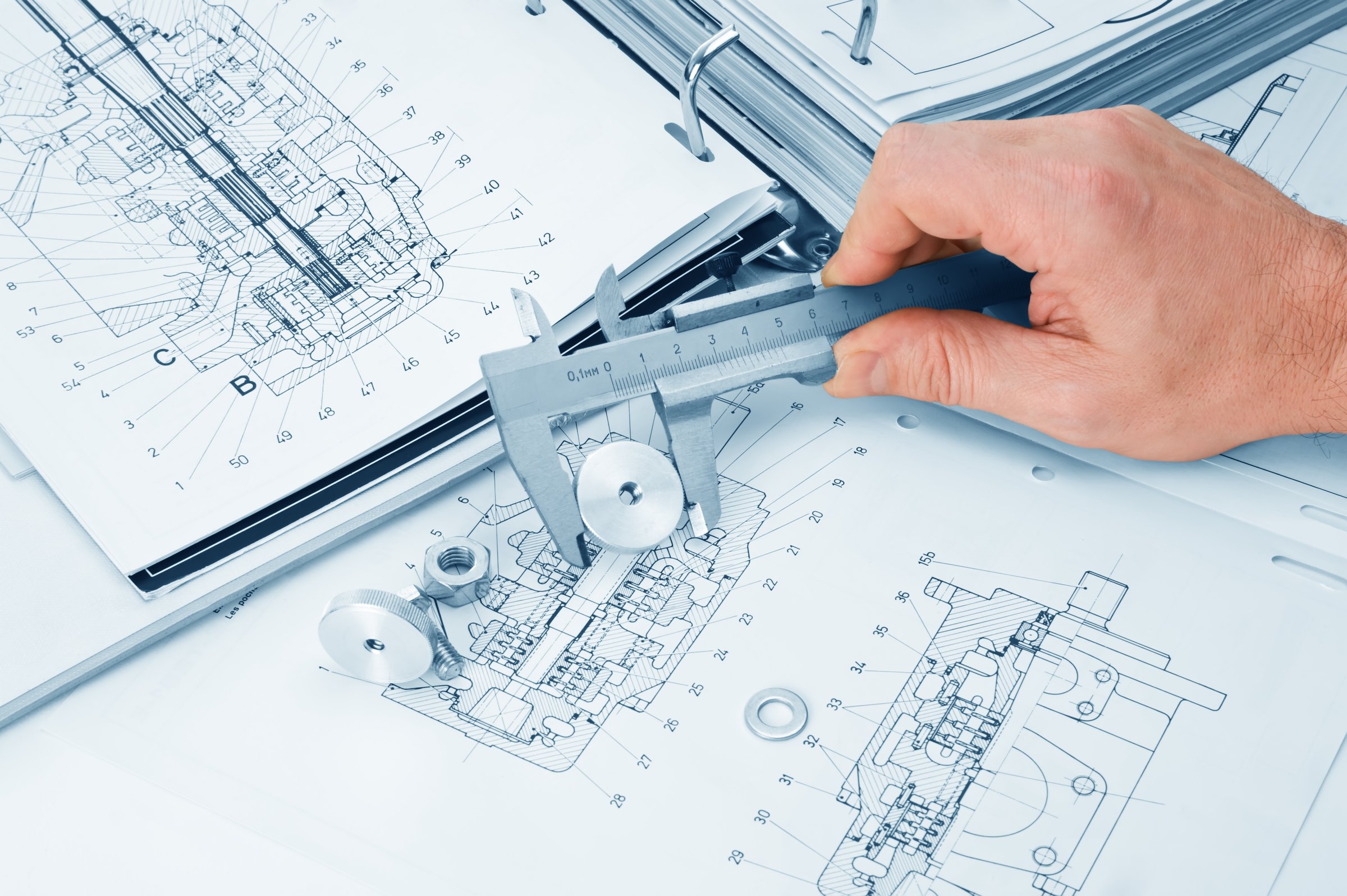 Person measuring a metal component with calipers on mechanical engineering blueprints.