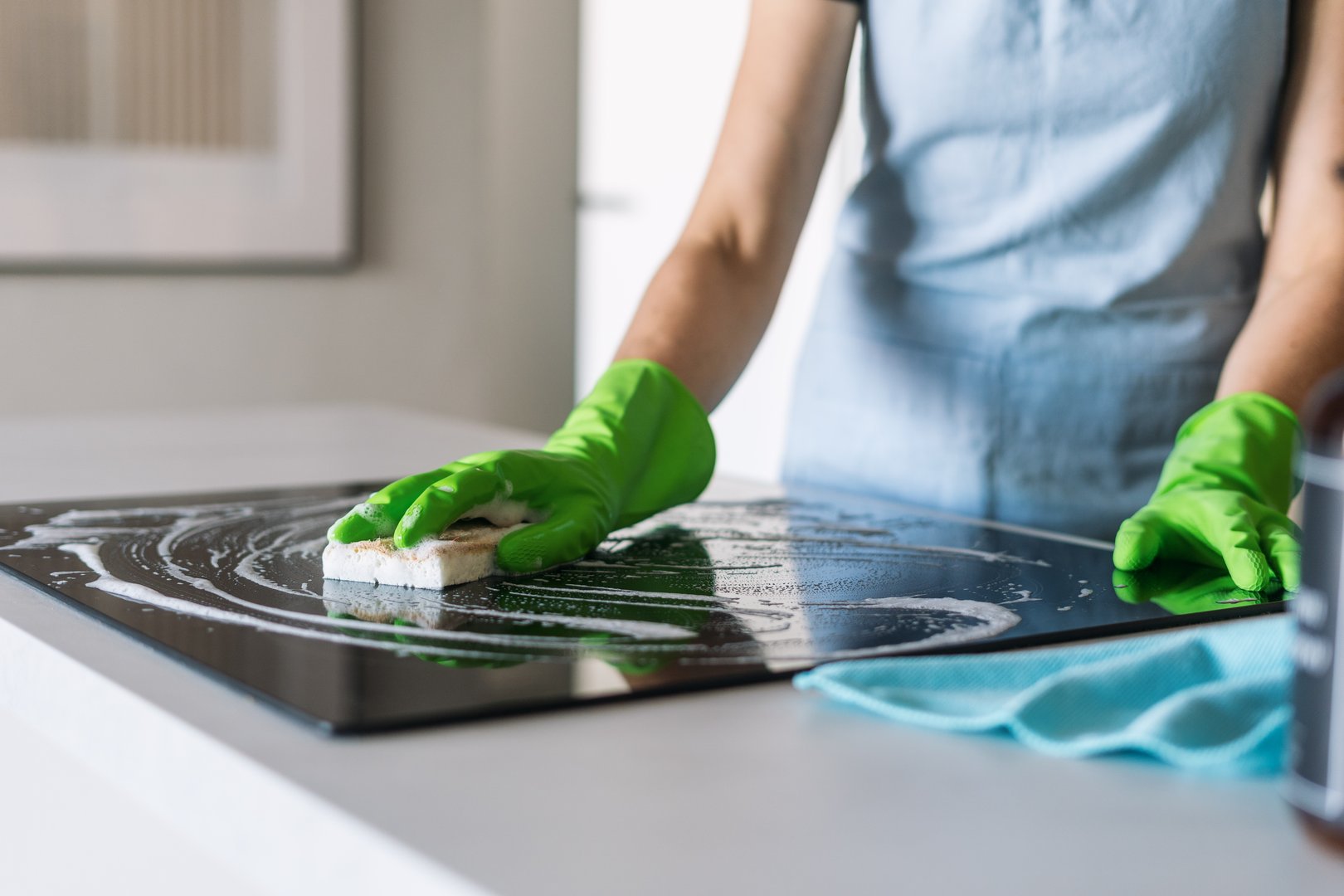 A woman in green gloves cleans a sleek induction stove with a sponge, maintaining the polished surface of her modern kitchen countertop during her housekeeping routine.